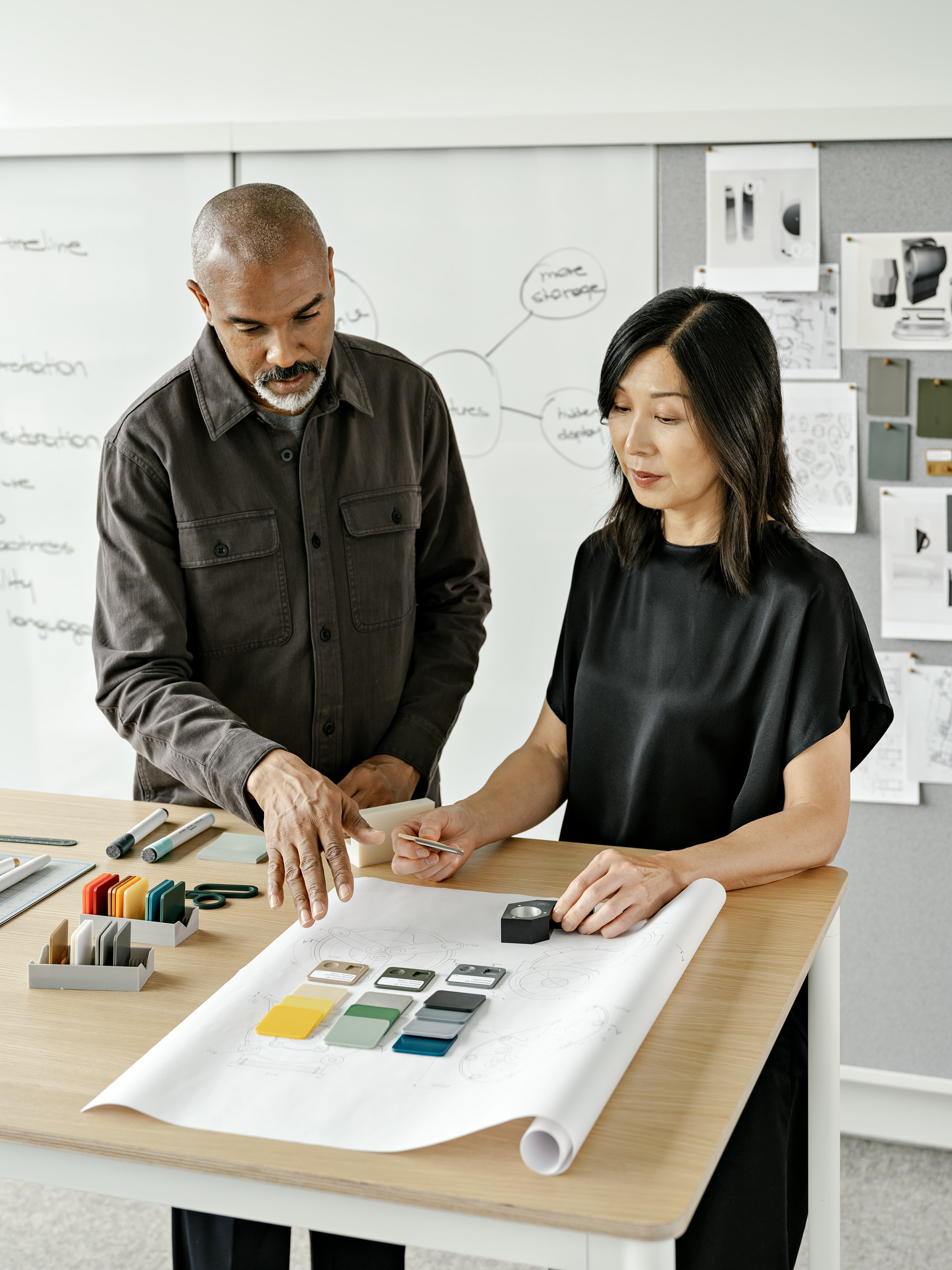 Two people standing at a table and working on a design sketch. Several color and material samples can be seen placed about the table.