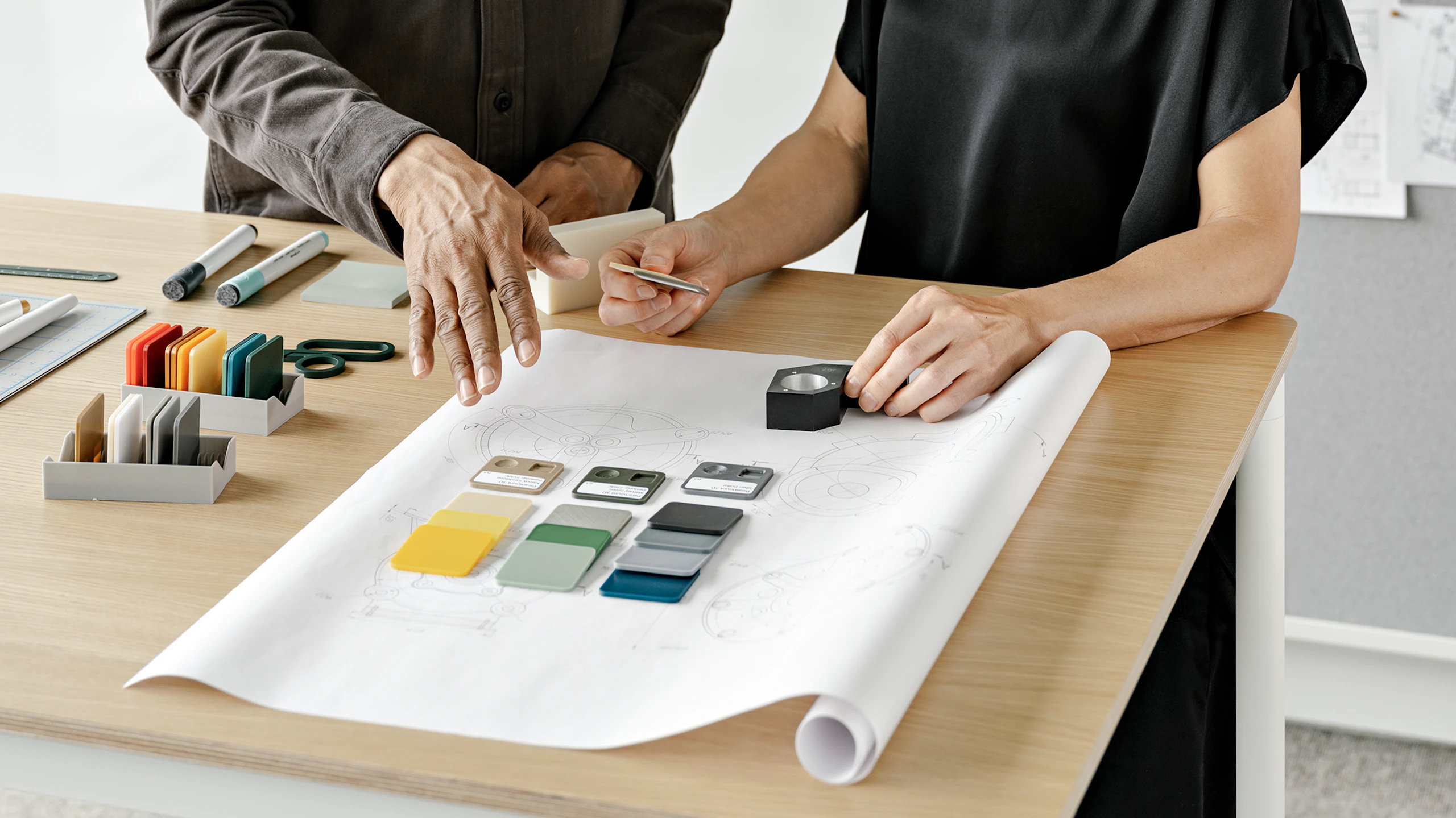 Two people standing at a table and working on a design sketch. Several color and material samples can be seen placed about the table.