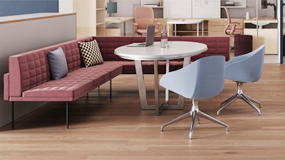 Zona de reuniones con un sofá acolchado rosa, una mesa redonda blanca y dos sillas de color azul claro. Sobre la mesa hay una computadora portátil y un vaso de agua. Estaciones de trabajo visibles en el fondo.