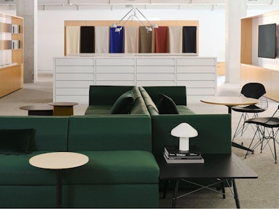 Several large green sofas and black side tables with a large showroom with textiles in a variety of colors lining the walls in the background at the MillerKnoll showroom at Design Days