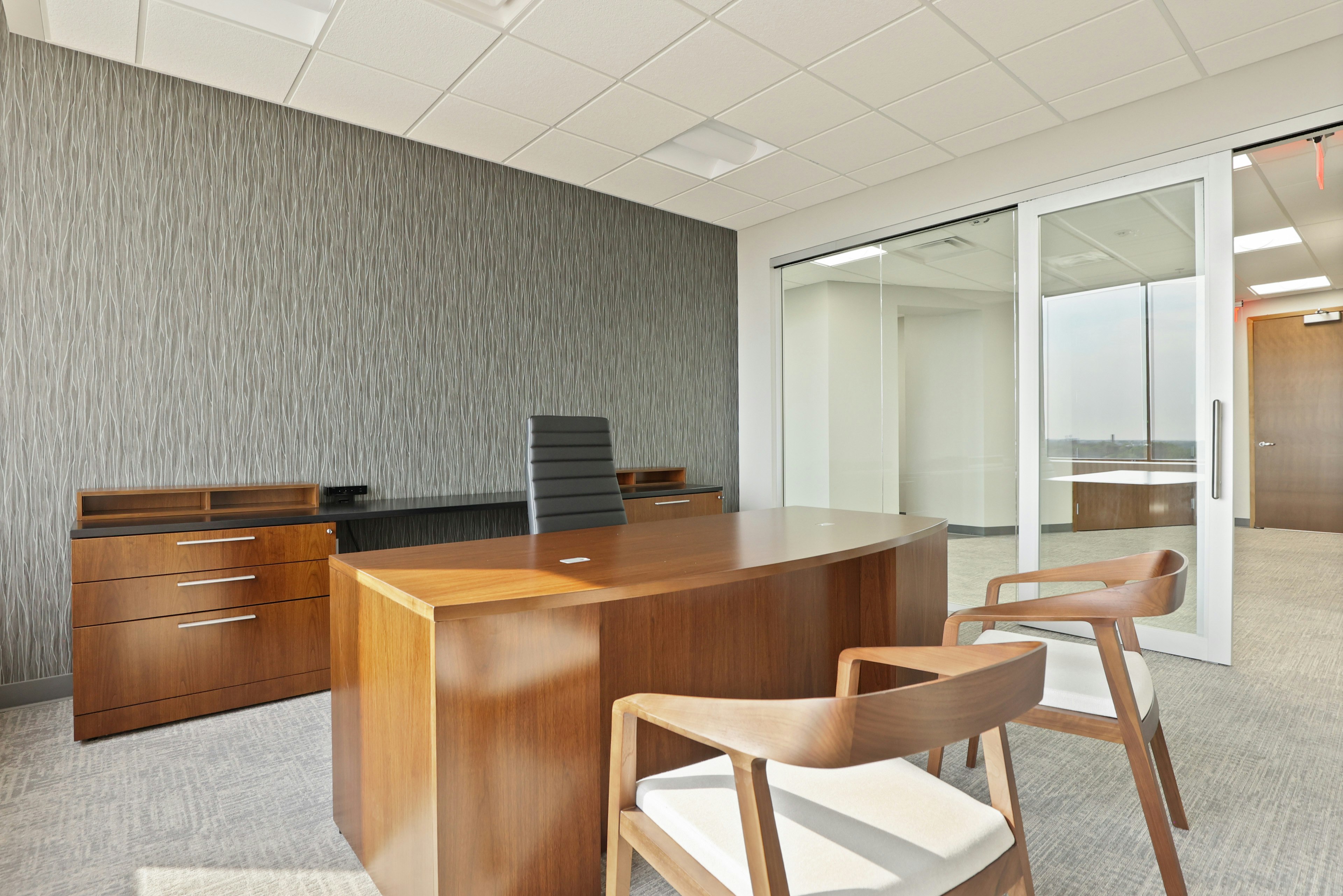 A private office setting at the Genesee County administration building furnished with a large wooden desk, a black executive chair, and two wooden chairs with white cushions.