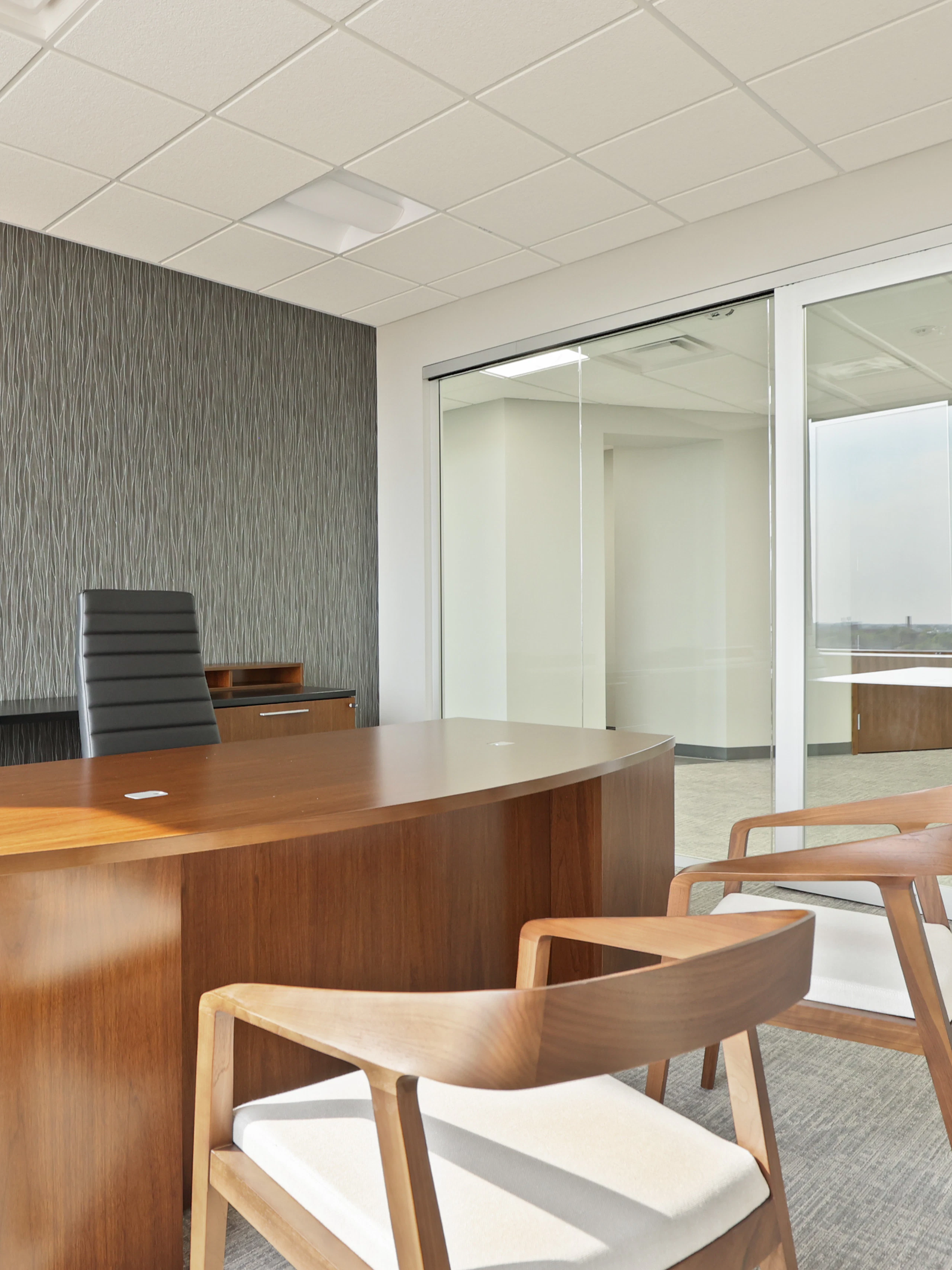 A private office setting at the Genesee County administration building furnished with a large wooden desk, a black executive chair, and two wooden chairs with white cushions.