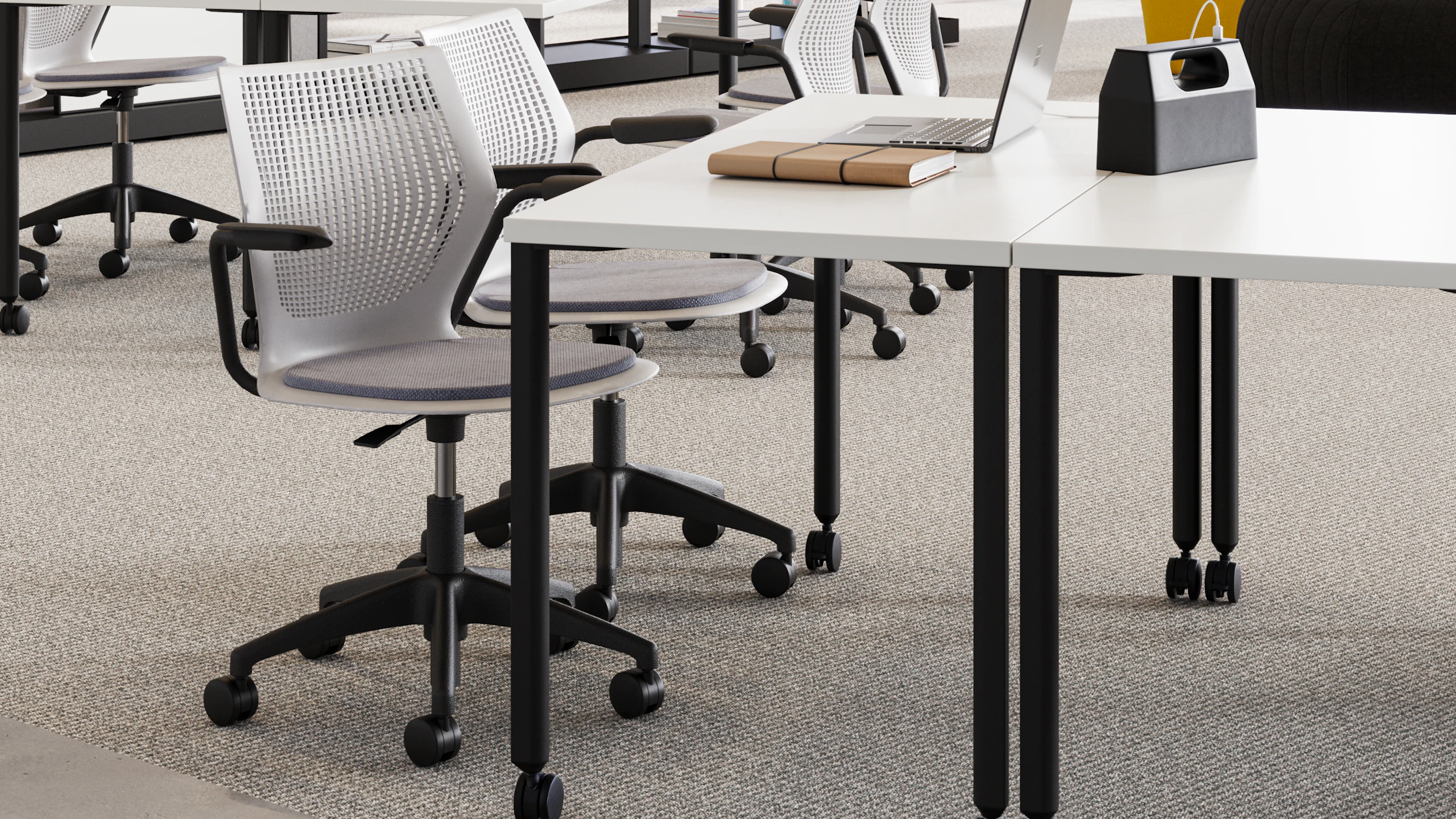 A laptop and book sitting on a black and white table that features white Herman Miller Fuld Nesting chairs dark blue seats next to a Herman Miller OE1 storage trolley