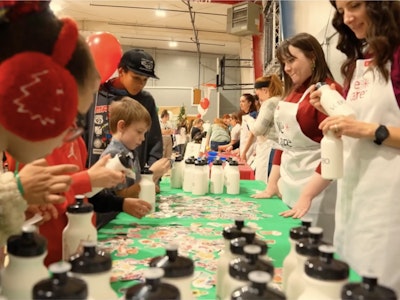A table of volunteers at a We Care event talking with several children. Water bottles and stickers can be seen on a green table.
