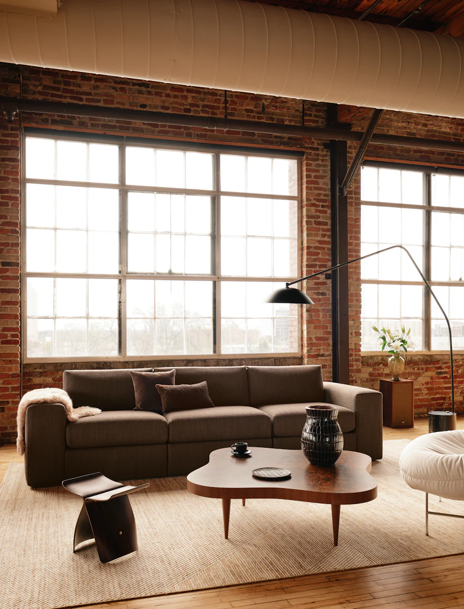 Living room scene featuring a gray sofa set, a coffee table, and a curved black floor lamp in a brick loft.