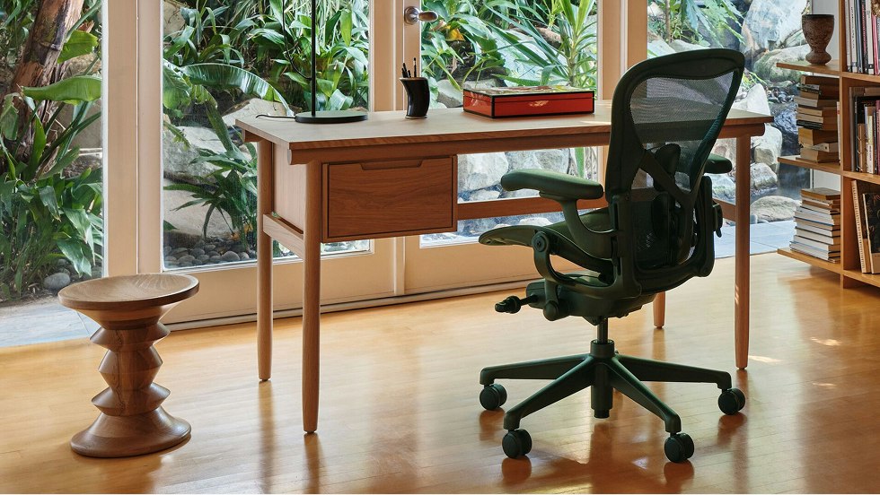 A Raleigh Desk from DWR, black Aeron Chair from Herman Miller, and Eames Turned Stool next to large windows overlooking green foliage.