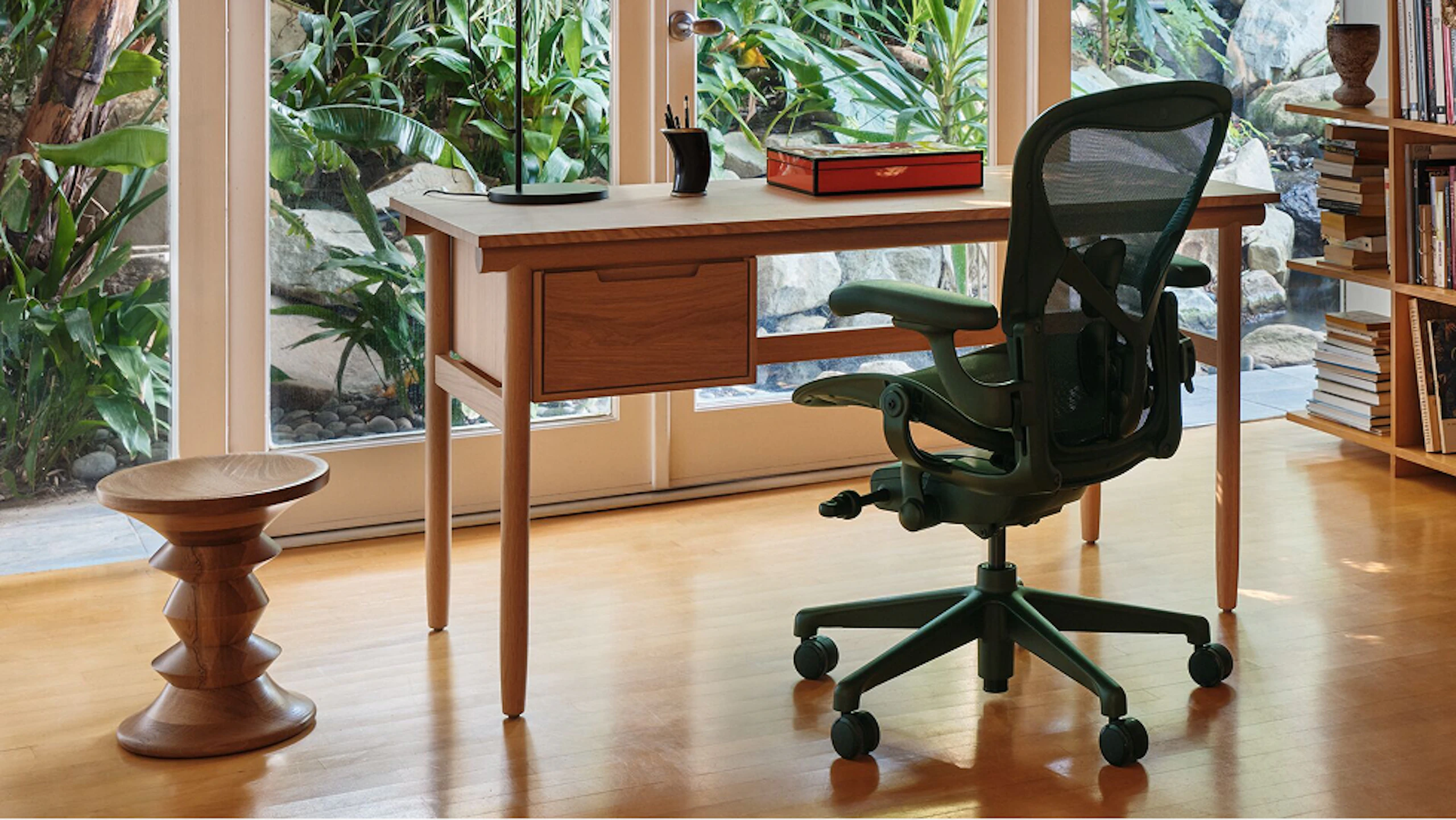 A Raleigh Desk from DWR, black Aeron Chair from Herman Miller, and Eames Turned Stool next to large windows overlooking green foliage.