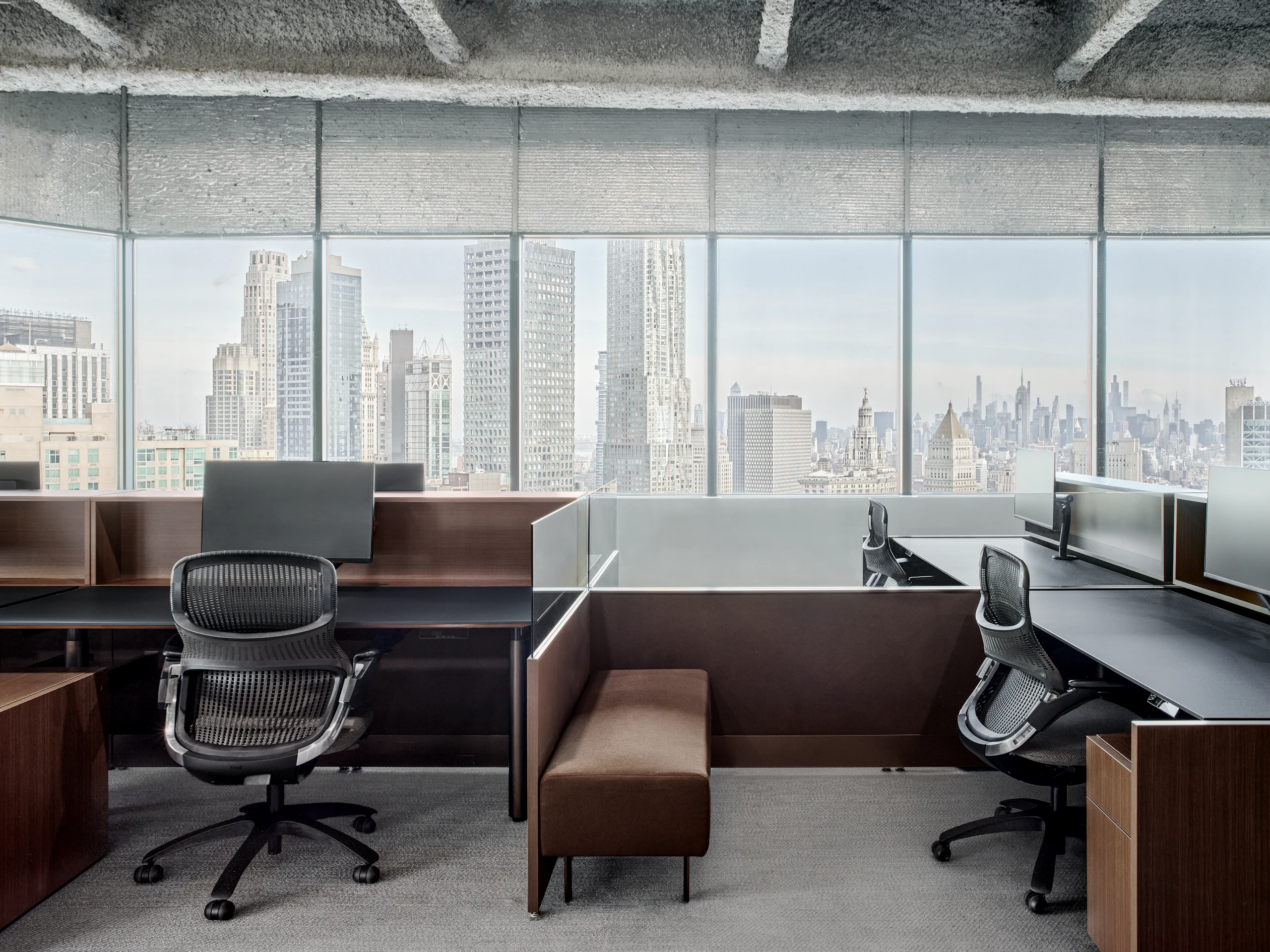 A group of workstations in front of a window with the New York City skyline outside. The workstations have a dark finish, bench seating for guests as well as a work chair and personal storage at each station.