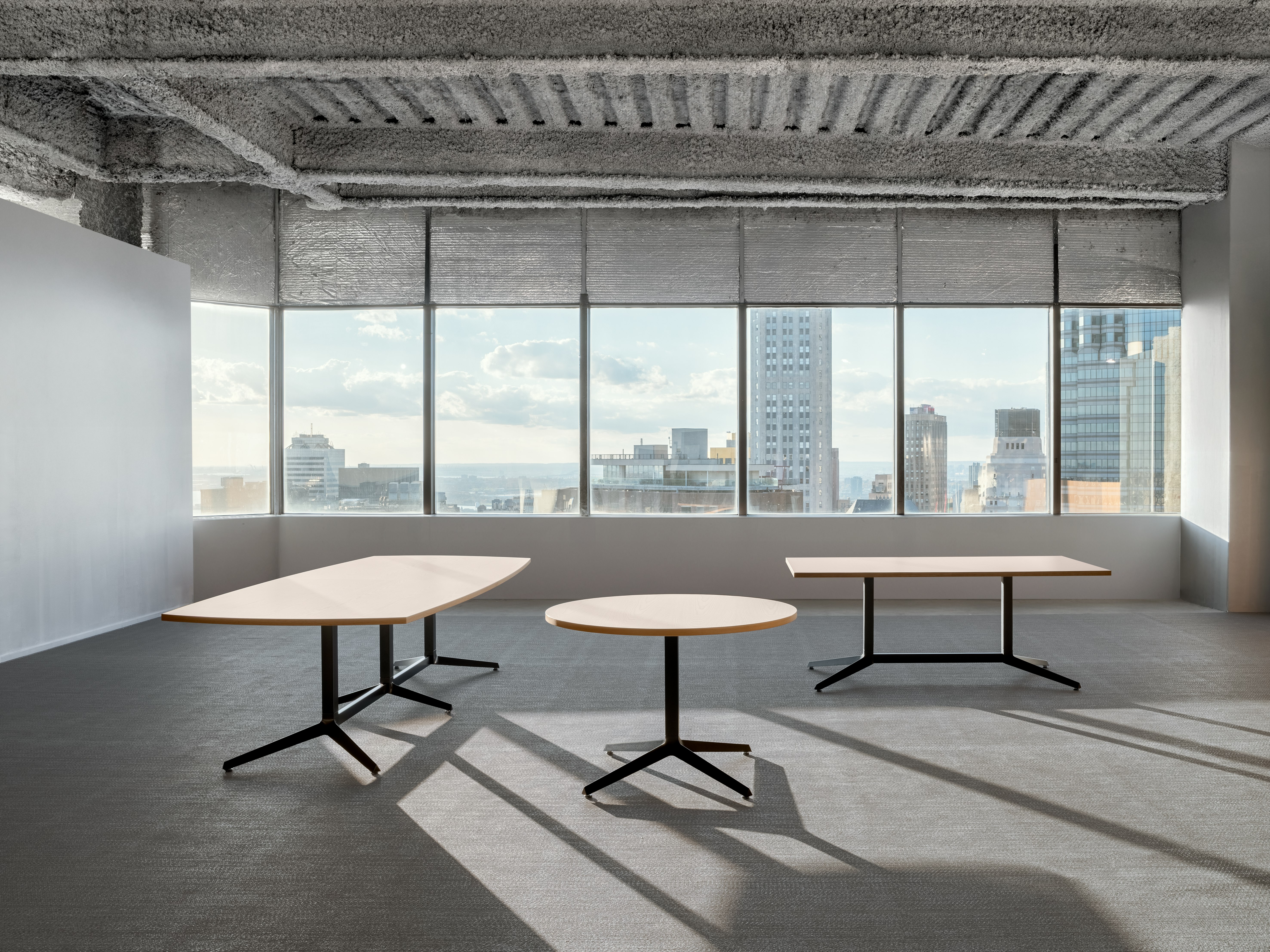 Three tables of various sizes and shapes are in an open office room in front of a large wall of windows.