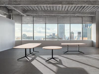 Three tables of various sizes and shapes are in an open office room in front of a large wall of windows.