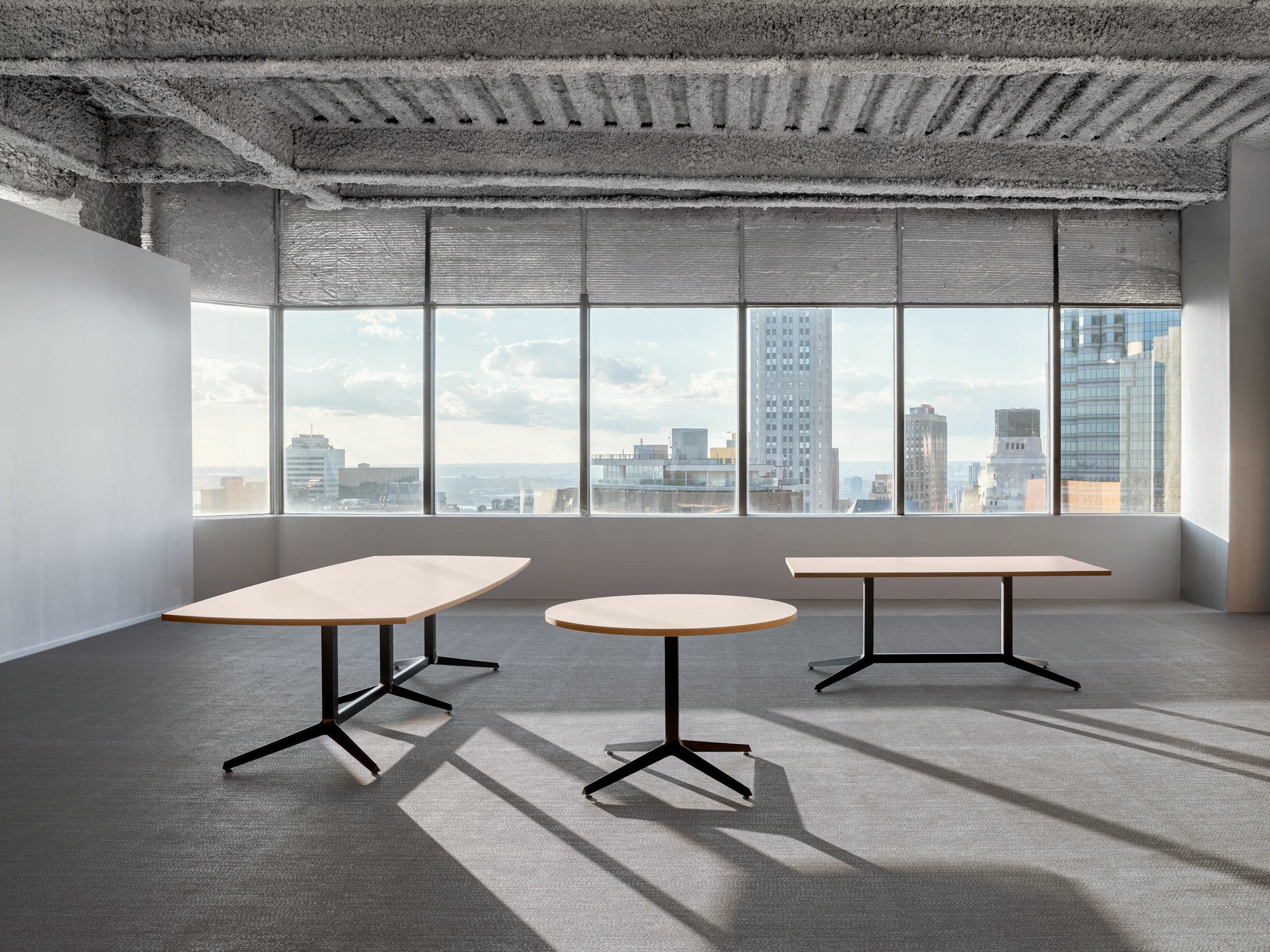 Three tables of various sizes and shapes are in an open office room in front of a large wall of windows.