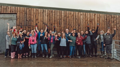 A group of people waving and posing for a photo in front of a building with a wall made of wooden panels.