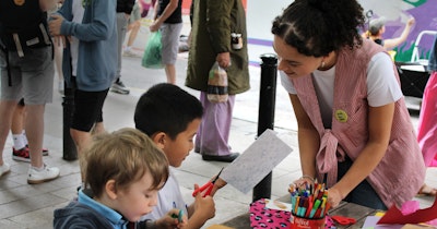 A woman wearing a red and white striped vest helping two children do arts and crafts. Several people can be seen standing on the sidewalk in the background.