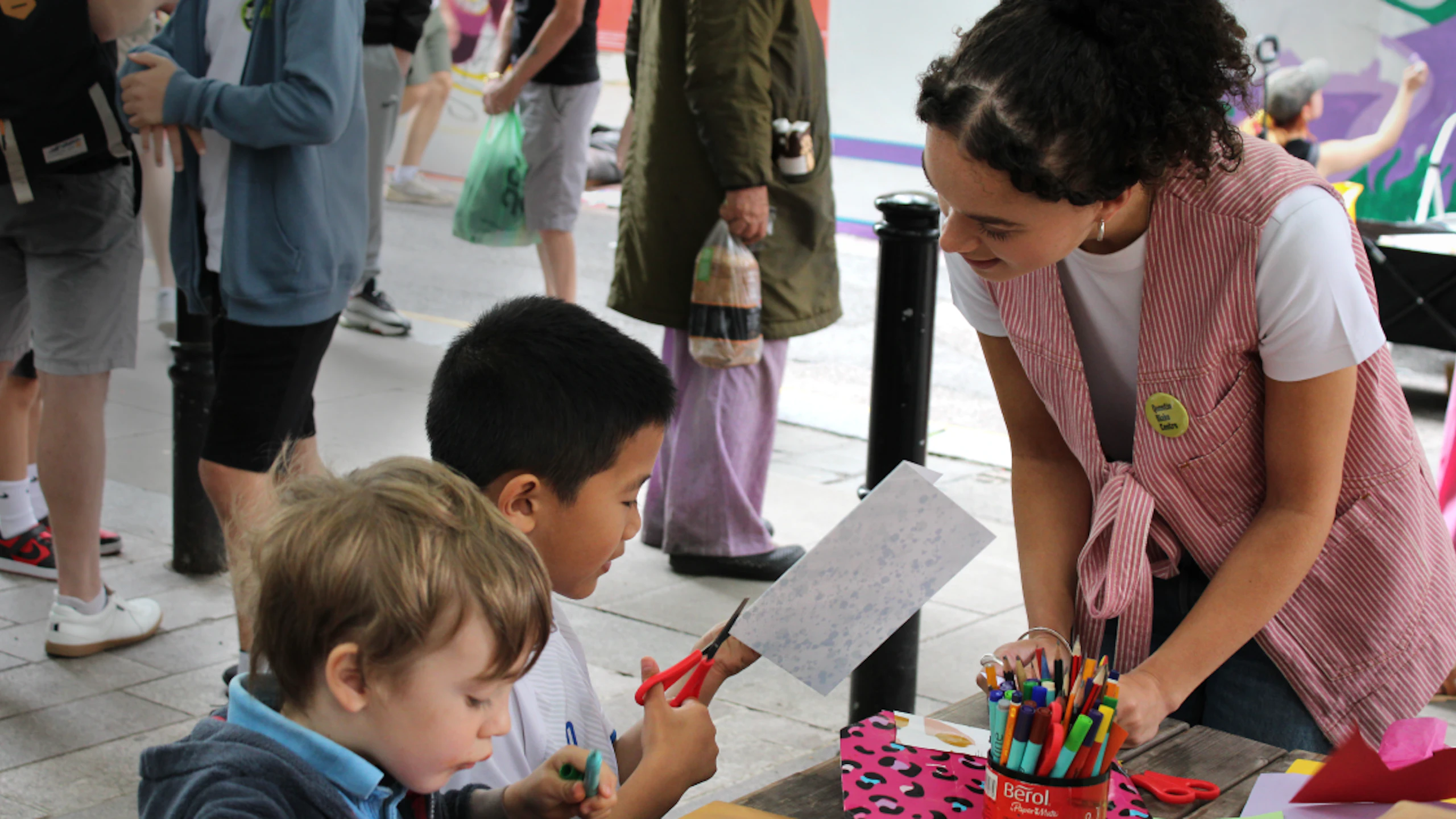 A woman wearing a red and white striped vest helping two children do arts and crafts. Several people can be seen standing on the sidewalk in the background.