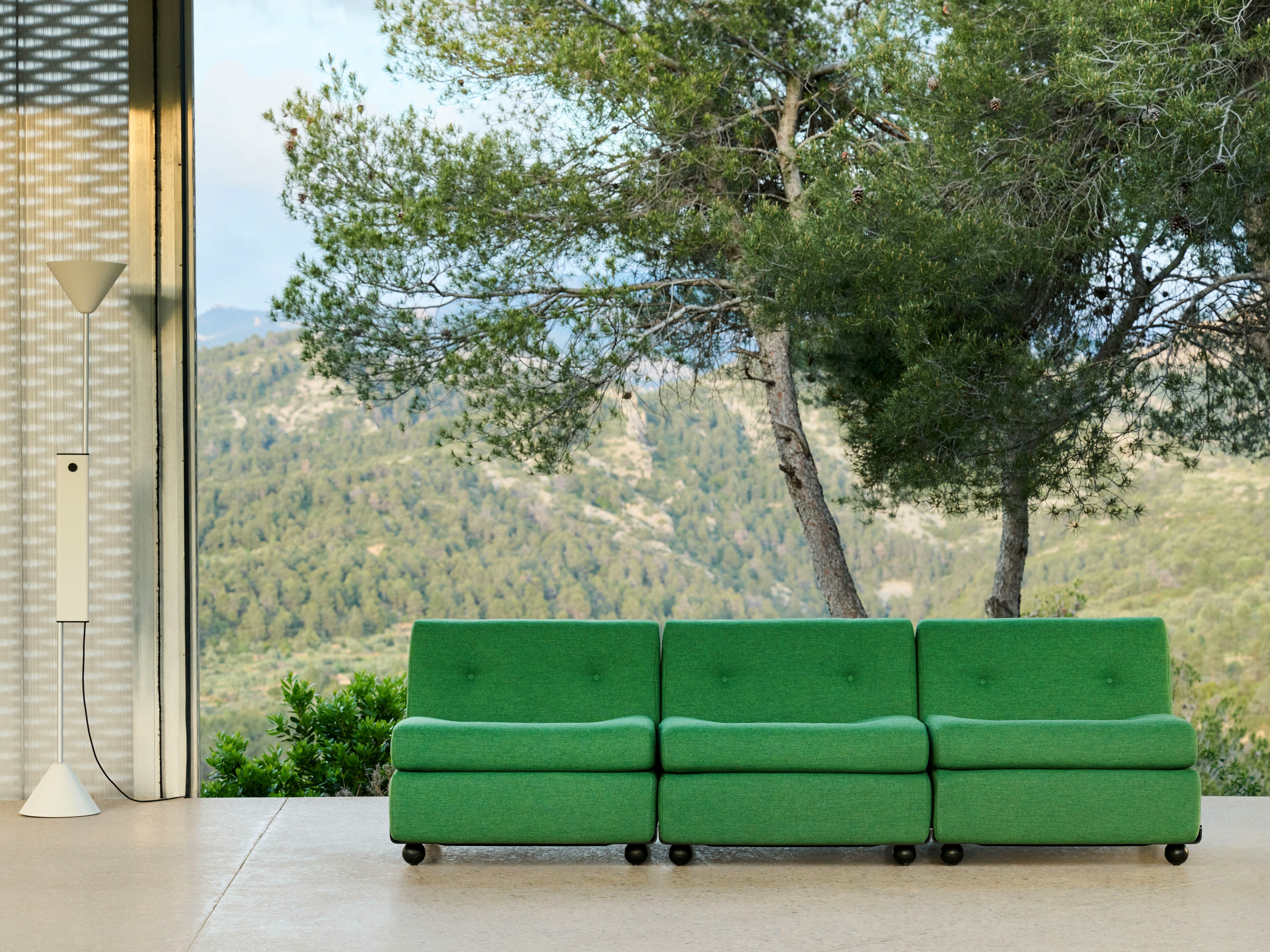 Three-seat green upholstered Amanta sofa in outdoor setting with tree and concrete wall backdrop.
