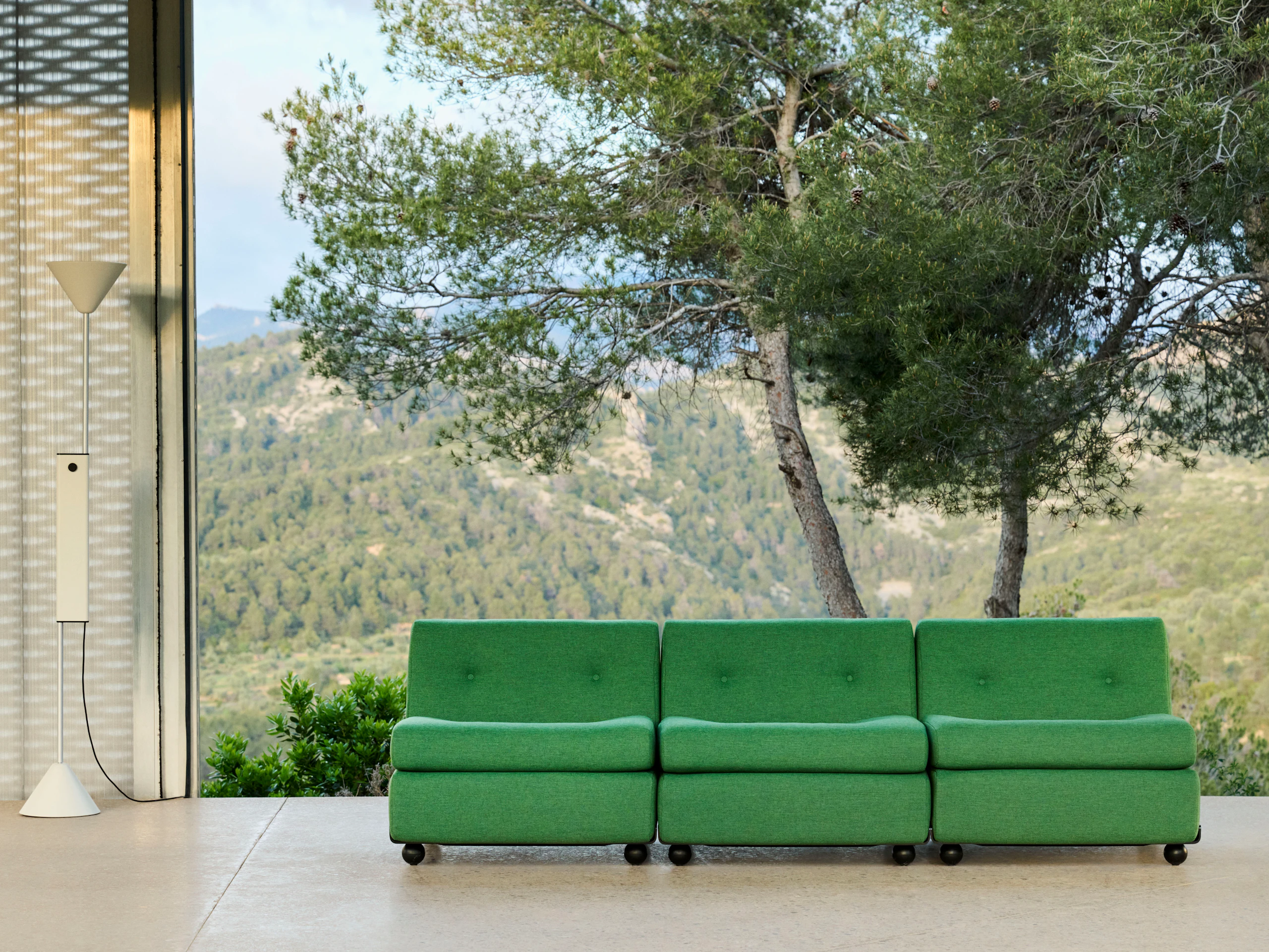 Three-seat green upholstered Amanta sofa in outdoor setting with tree and concrete wall backdrop.