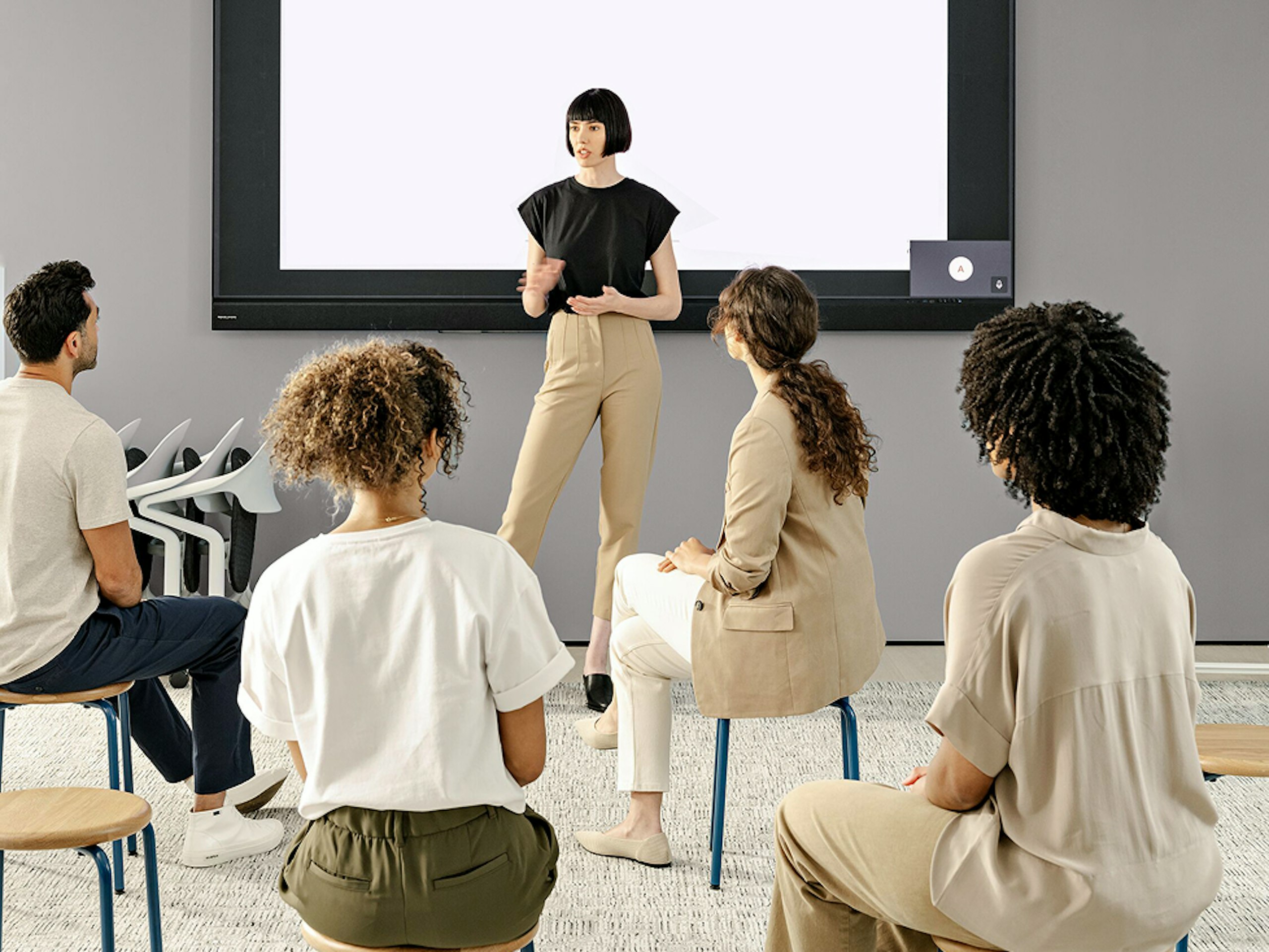 A woman standing in front of a screen giving a presentation to an audience of three women and one man seated on stools.