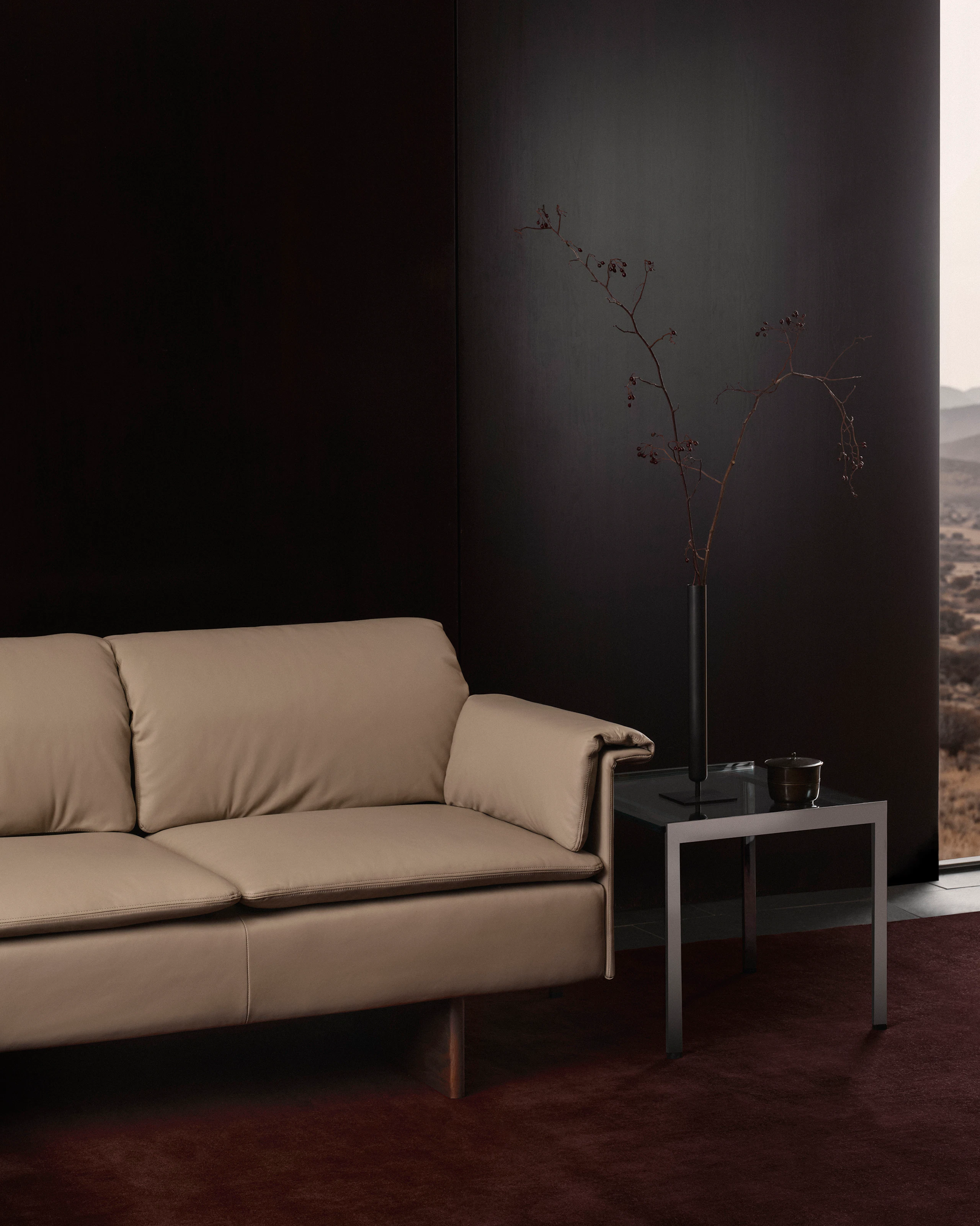 Mantle Sofa designed by Ward Bennett upholstered with Bristol Leather in Champagne beside an H Frame side table, viewed at an angle against a black wall on a burgundy rug.