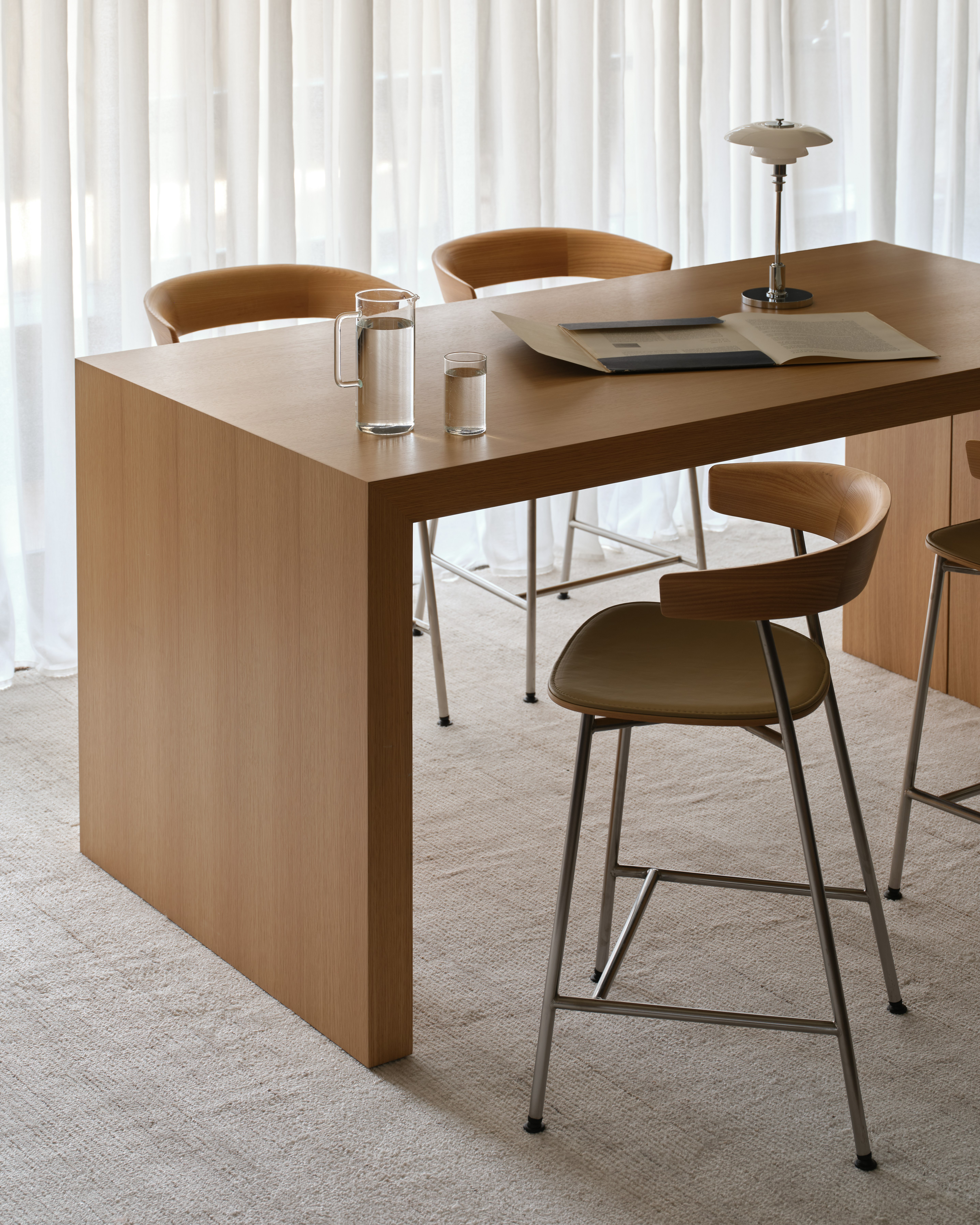 JD Waterfall Table in Natural Rift Cut Oak paired with four counter-height Leeway Stools in Oak with a satin chrome base, viewed at an angle on a beige rug against white curtains.