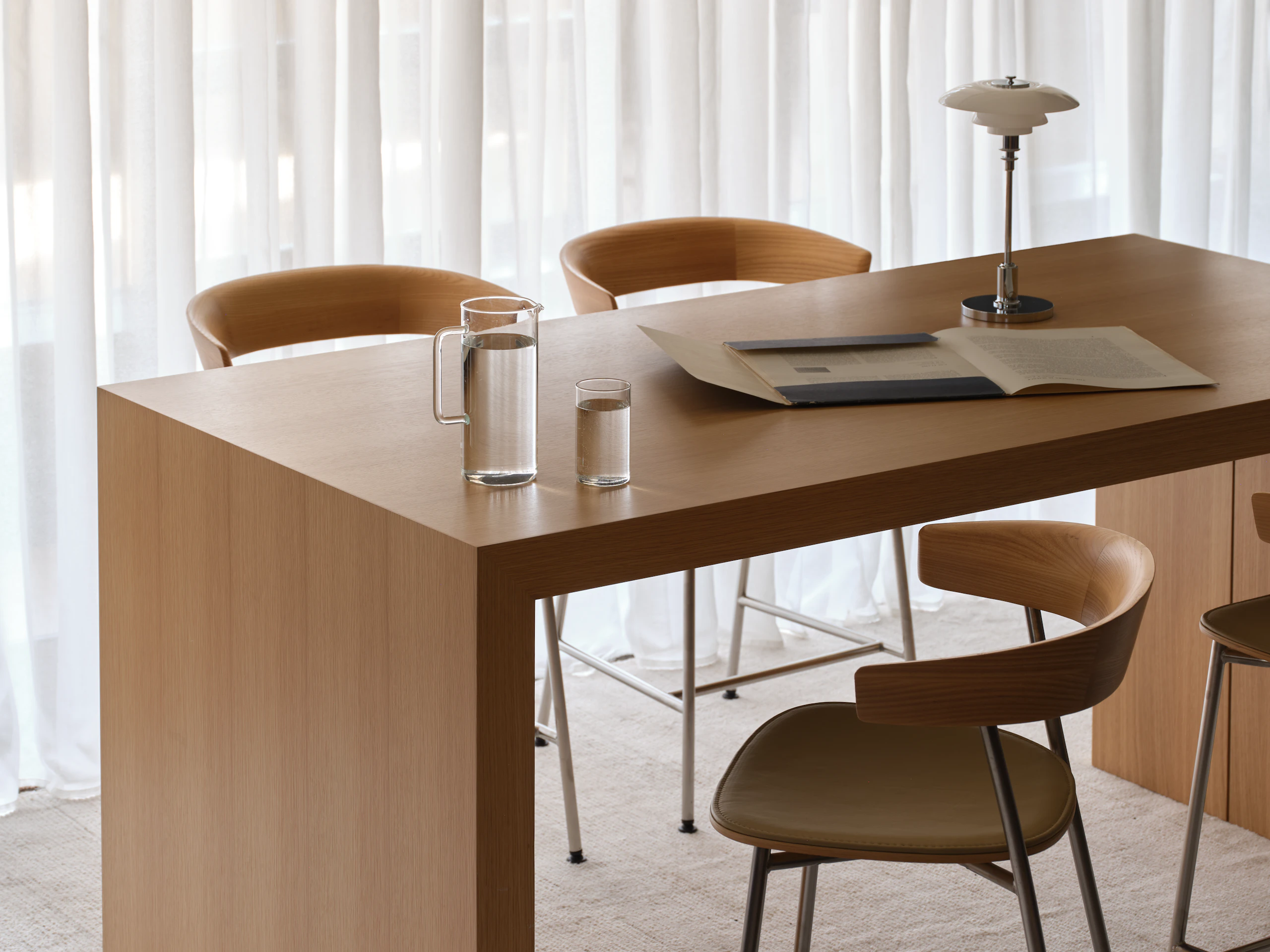 JD Waterfall Table in Natural Rift Cut Oak paired with four counter-height Leeway Stools in Oak with a satin chrome base, viewed at an angle on a beige rug against white curtains.