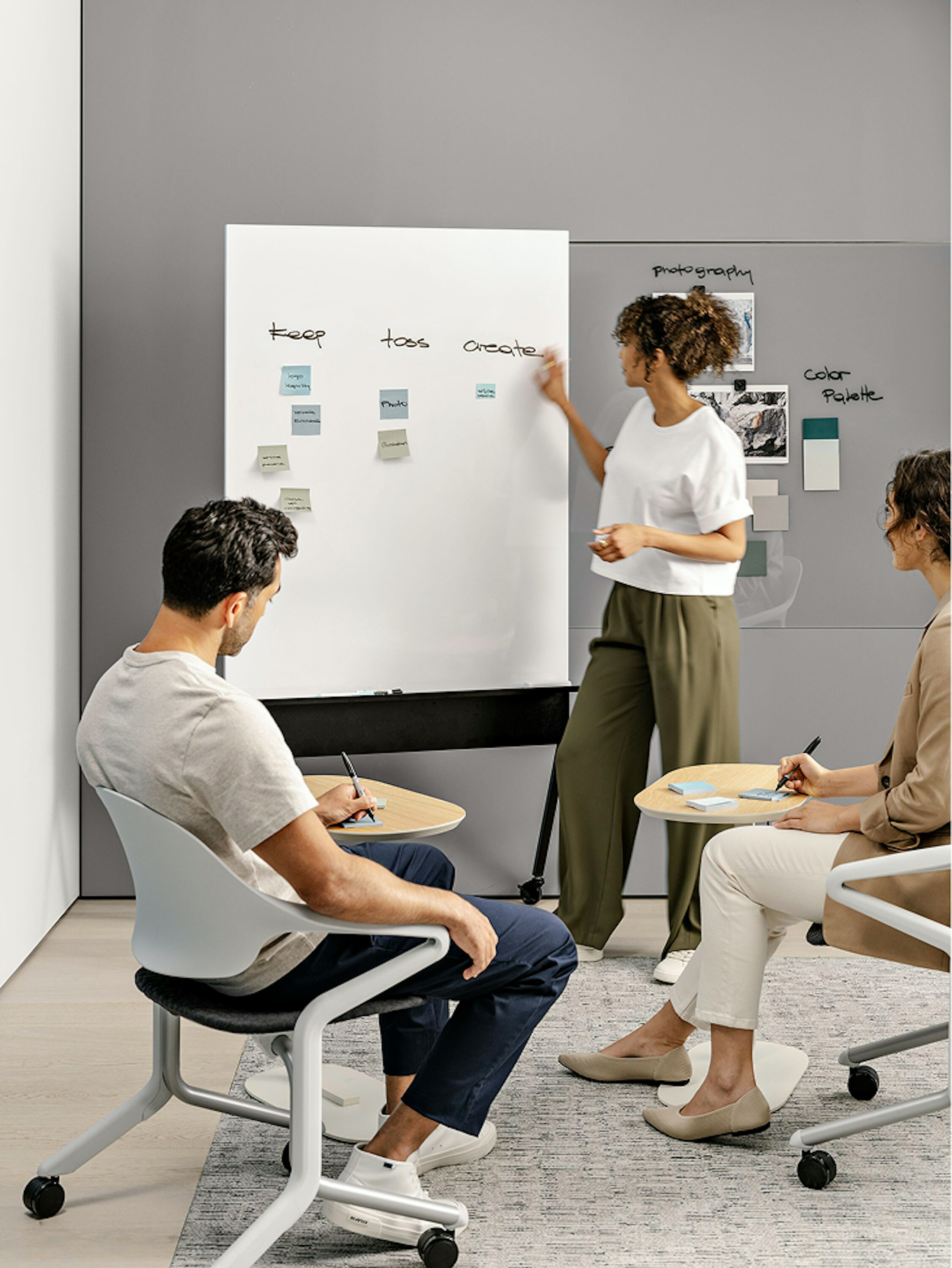 A person in a white shirt and green pants writing on a whiteboard while two people in black and white Herman Miller Fuld Nesting Chairs take notes.