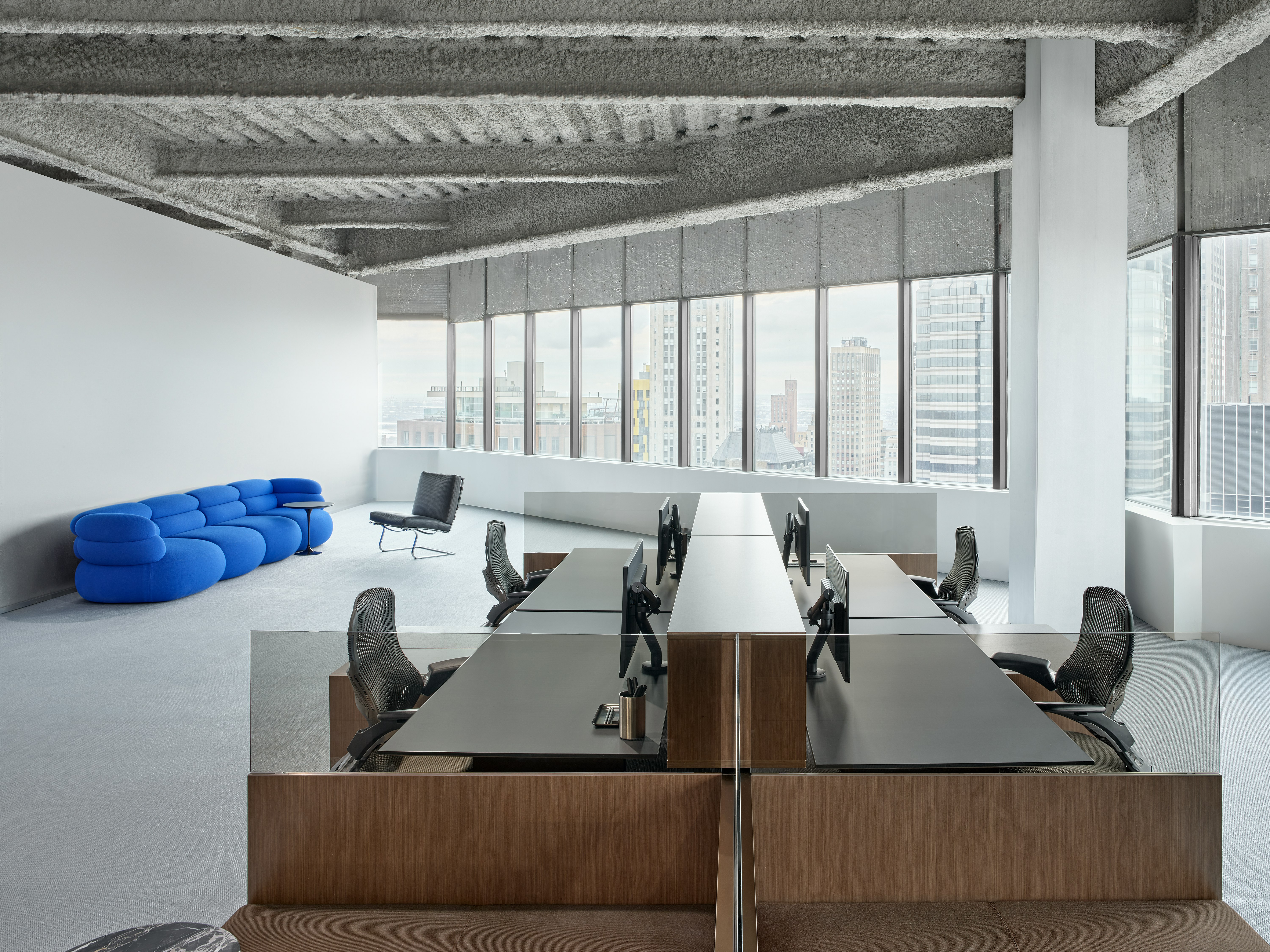 A group of four workstations are in front of a lounge seating area in front of a large row of windows in an open office.