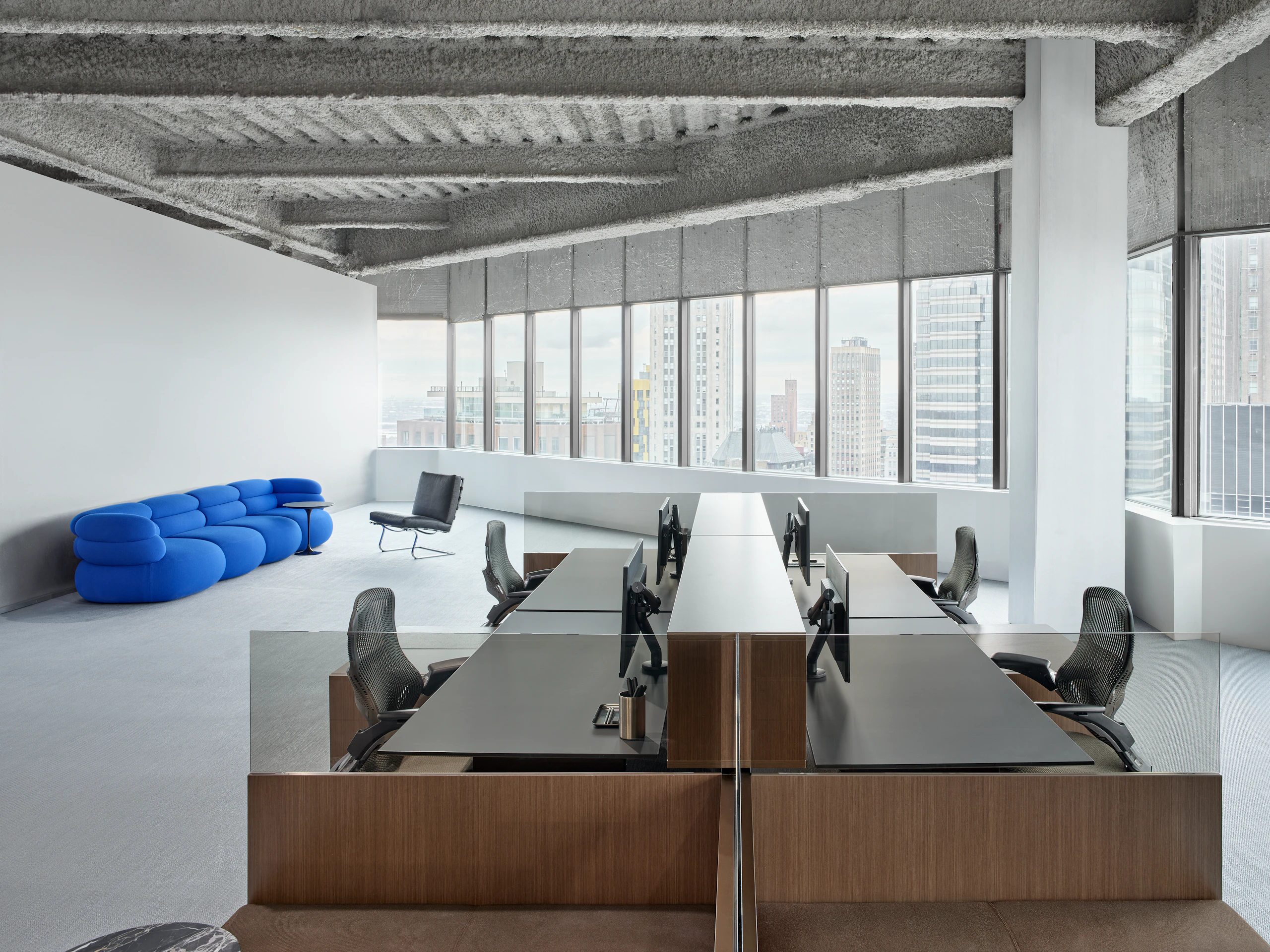 A group of four workstations are in front of a lounge seating area in front of a large row of windows in an open office.