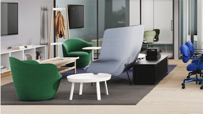 Moderna sala de estar con poltronas verdes y grises, una pequeña mesa blanca y un mueble de almacenaje negro. Cerca hay un perchero con chaquetas. Grandes ventanales y una oficina con paredes de cristal al fondo.