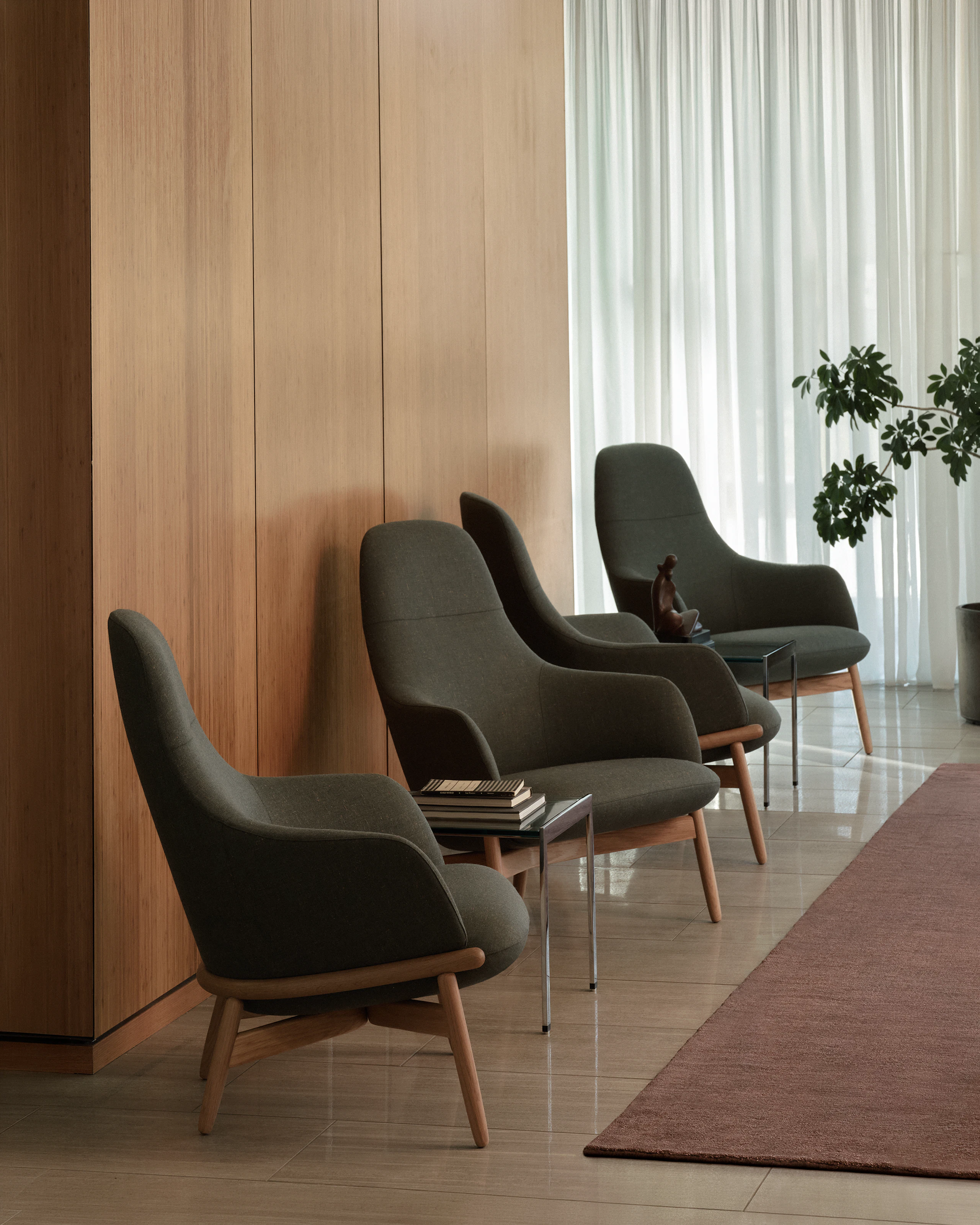 Four high-back Reframe Lounge Chairs in Oak upholstered with Rowan in Zucchini with two H Frame Side Tables, viewed at an angle against a wood wall.