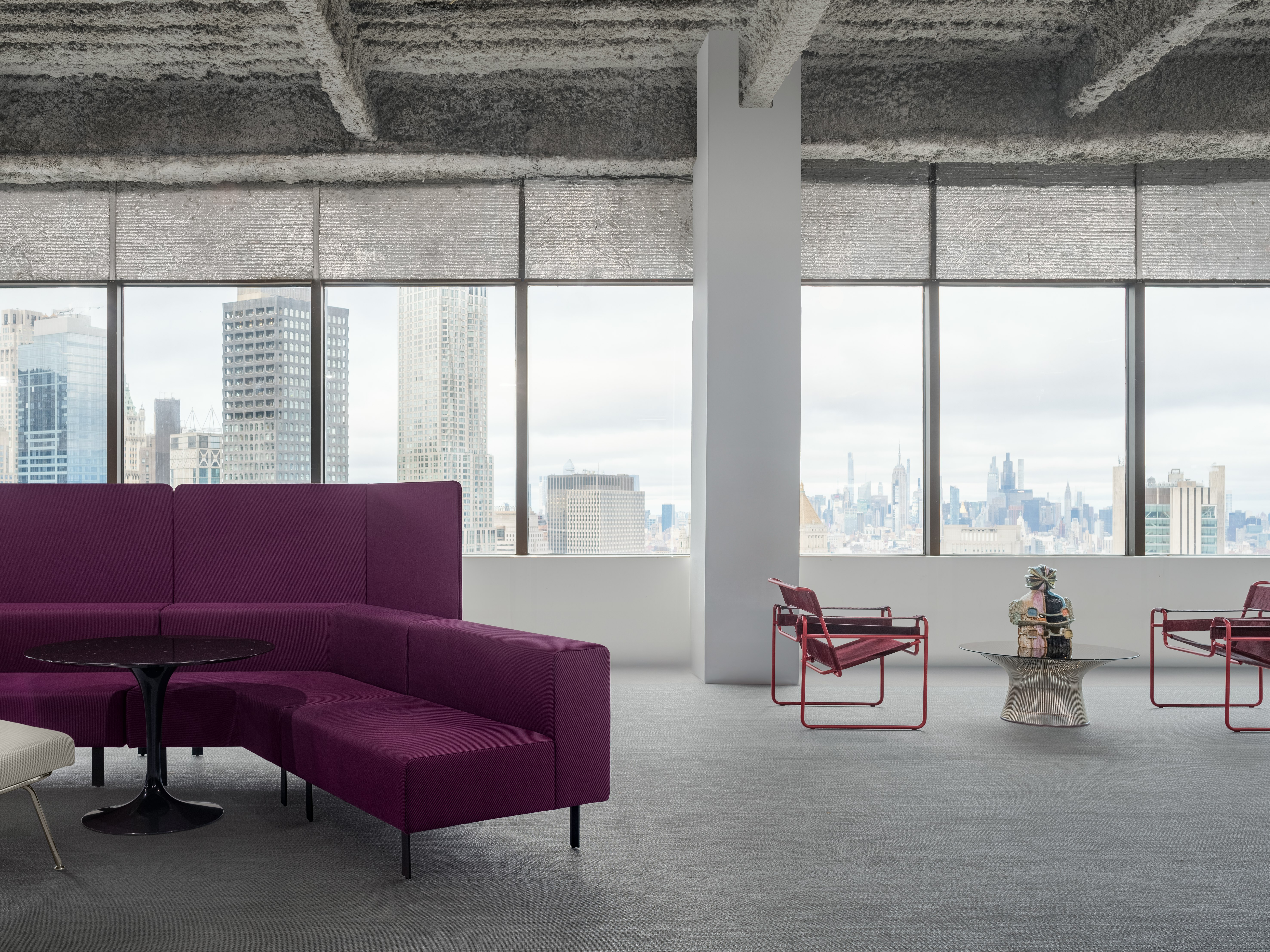 A lounge bench setting and a few lounge chairs with a coffee table are sitting in front of a long group of windows overlooking the New York City Skyline.