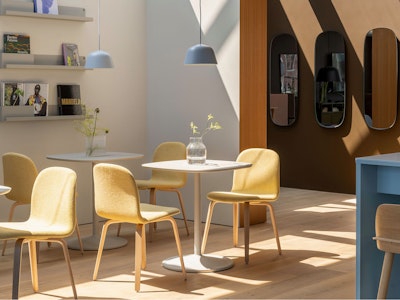 A brightly lit cafe setting furnished with Muuto furniture at MillerKnoll Design Days. Yellow chairs are arranged around small white tables with a row of wall mirrors in the background.