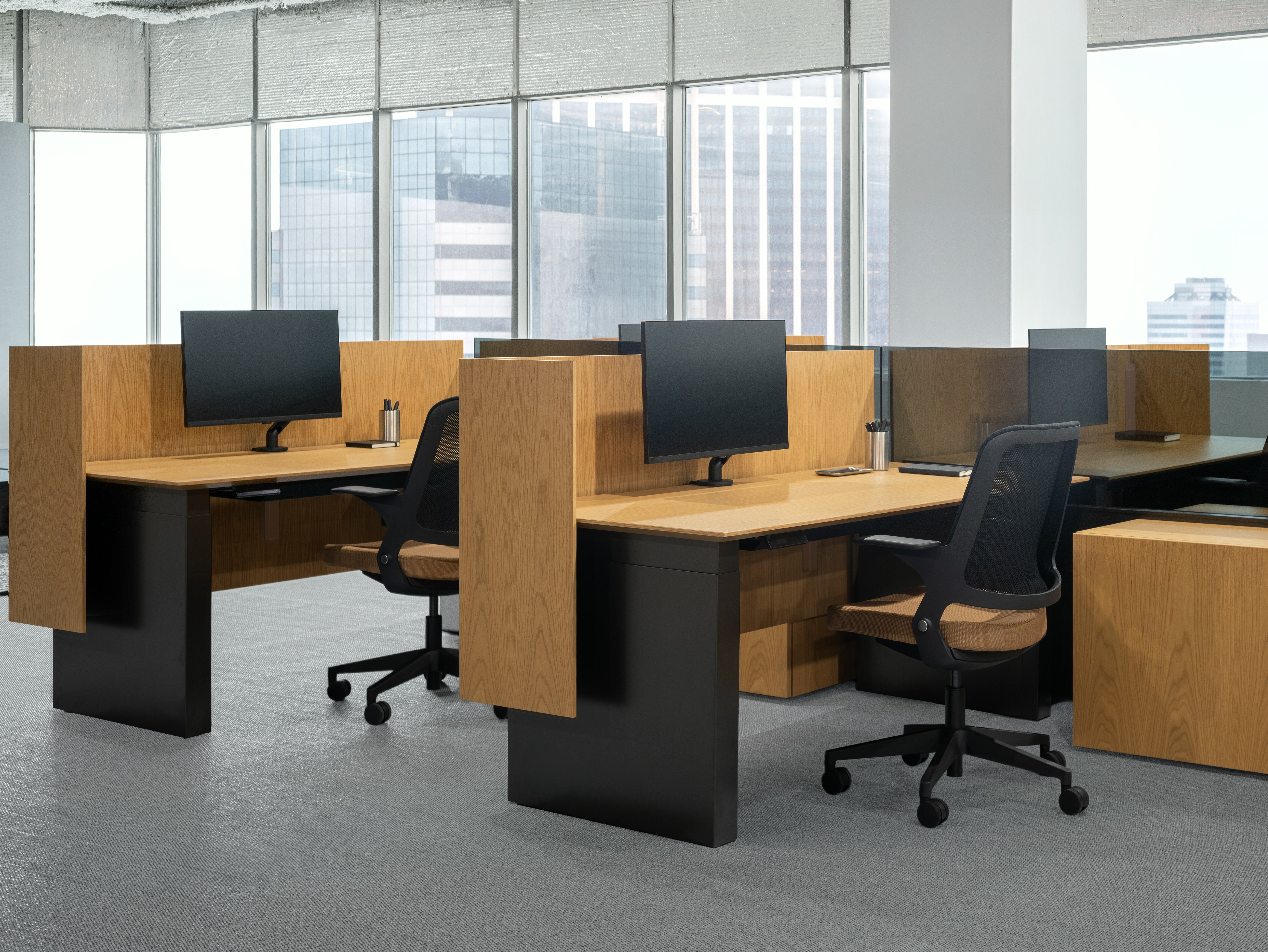 A group of panel based workstations are positioned in front of a large wall of windows. Each workstation is separated by screens and has a height adjustable table, storage credenzas and a work chair.