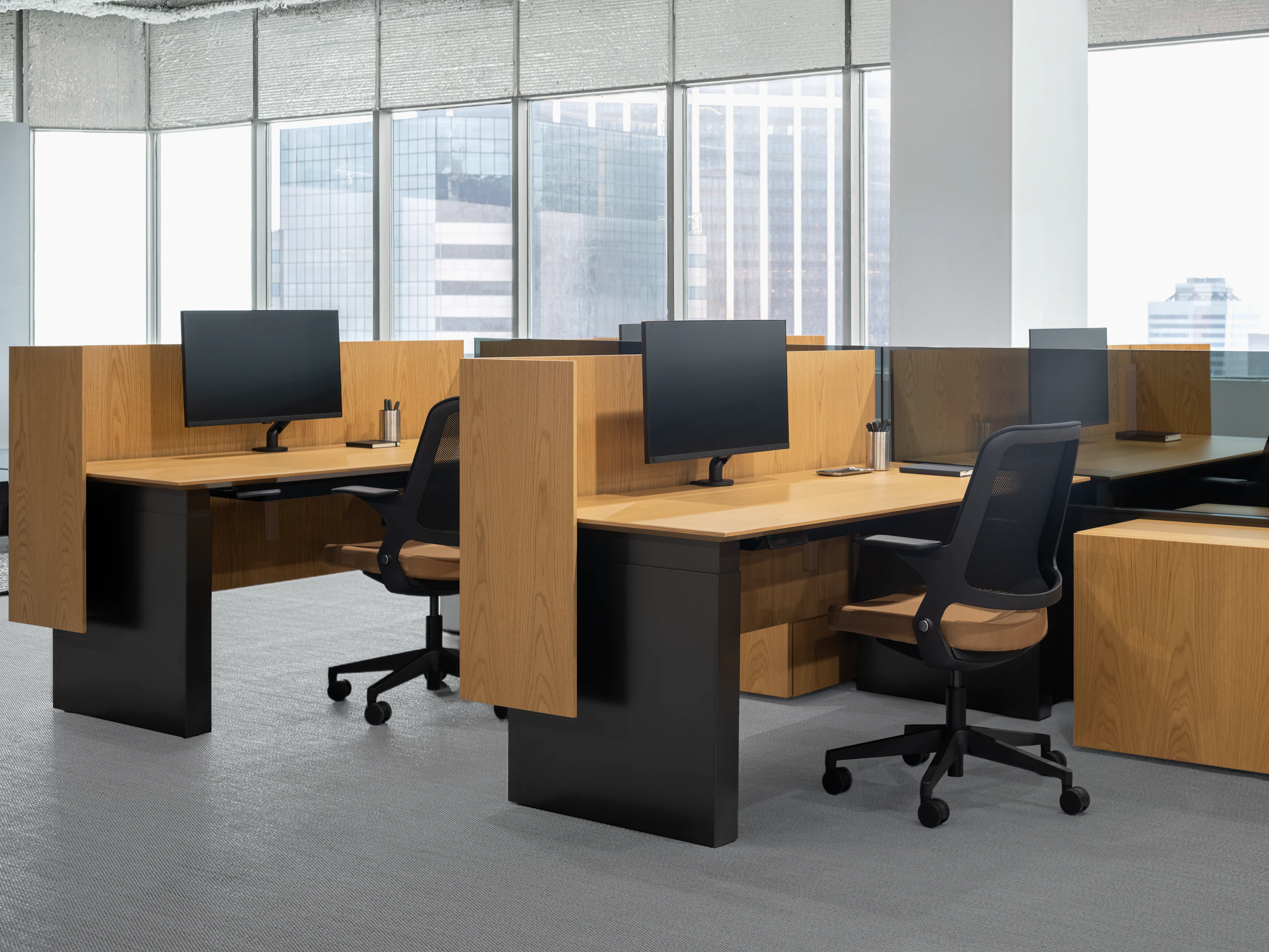 A group of panel based workstations are positioned in front of a large wall of windows. Each workstation is separated by screens and has a height adjustable table, storage credenzas and a work chair.
