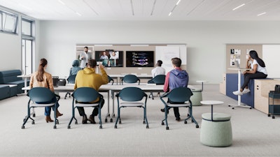 Aula moderna con foco en la colaboración. En primer plano se ve una larga mesa blanca con computadoras portátiles. El fondo muestra cabinas cerradas con paneles de tela roja y marcos blancos, algunas con asientos y pequeñas mesas en el interior.