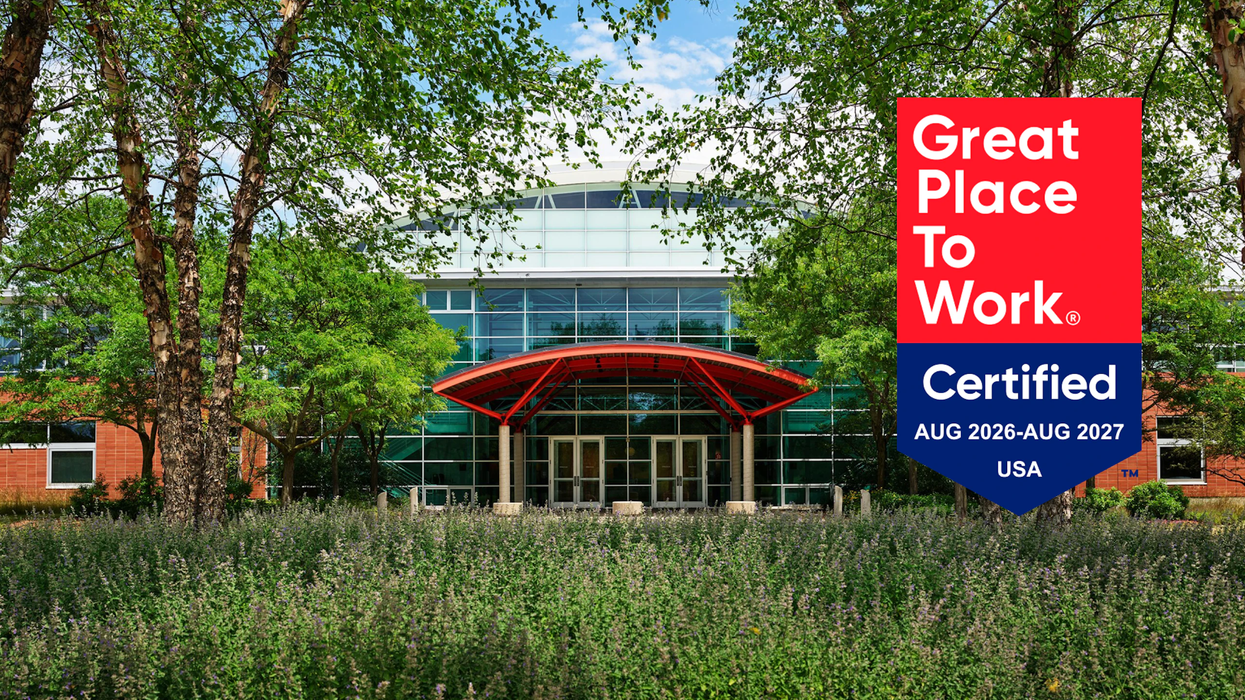 An exterior front view of the MillerKnoll Greenhouse manufacturing facility with the red and blue Great Place to Work certified 2026-2027 badge overlayed on top.