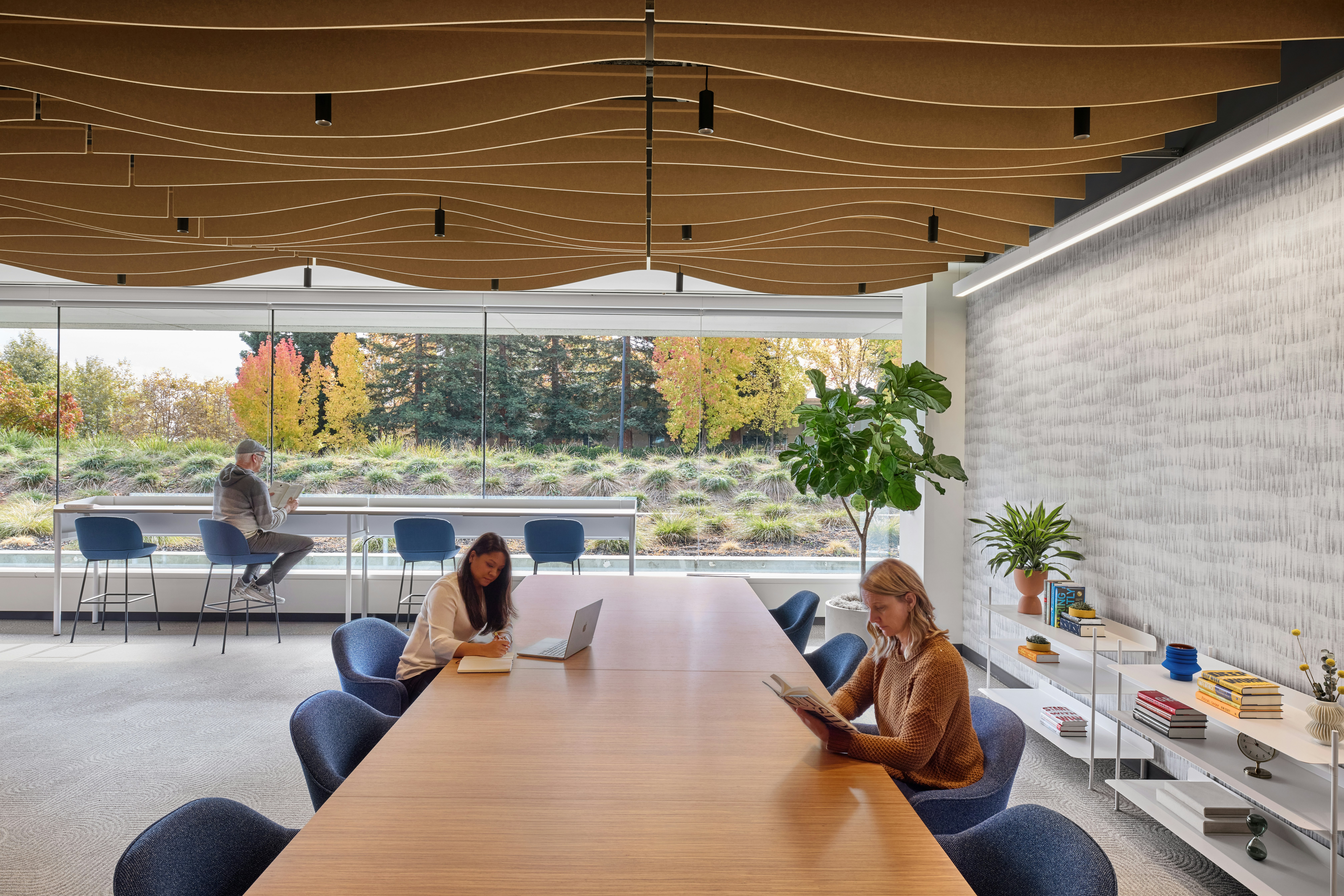 Shared work and social space with large communal table, ergonomic seating, elevated counter seating along windows, and sculpted acoustic ceiling elements.