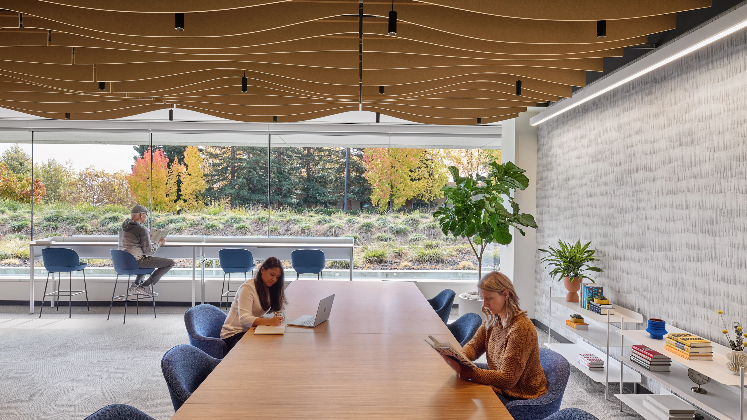 Shared work and social space with large communal table, ergonomic seating, elevated counter seating along windows, and sculpted acoustic ceiling elements.