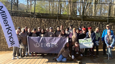 A group of people posing around a white flag featuring the MillerKnoll logo, with a banner displaying the Geiger logo next to it.