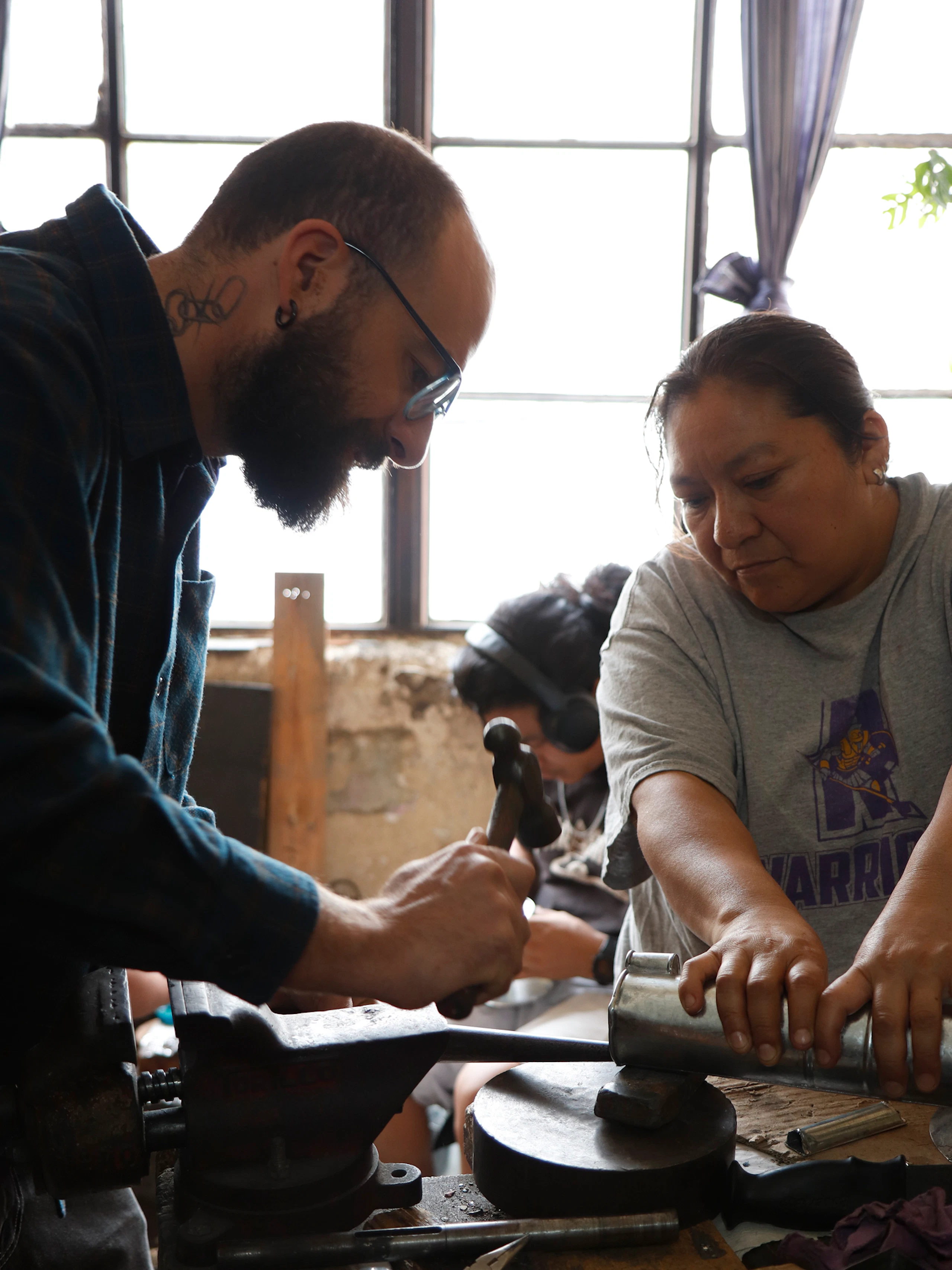 A man in glasses hammering a metal tube while a woman holds the tube in place on an anvil.