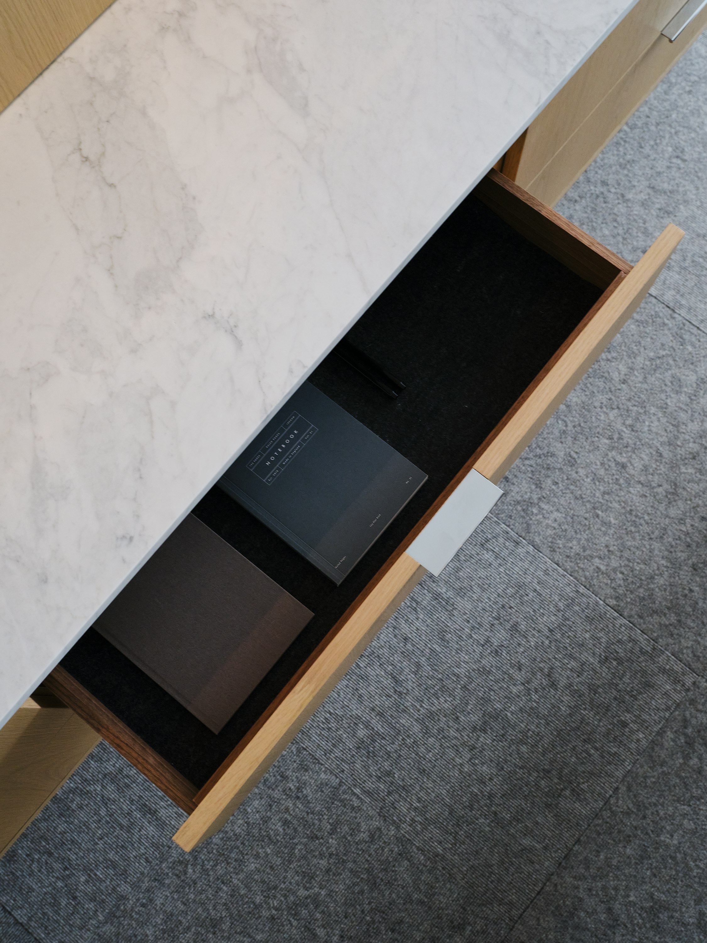 Detail view of a Geiger One Casegoods workwall open storage drawer in Nordic Flat Cut Oak with a White Carrara Marble top, viewed from above in a private office setting.