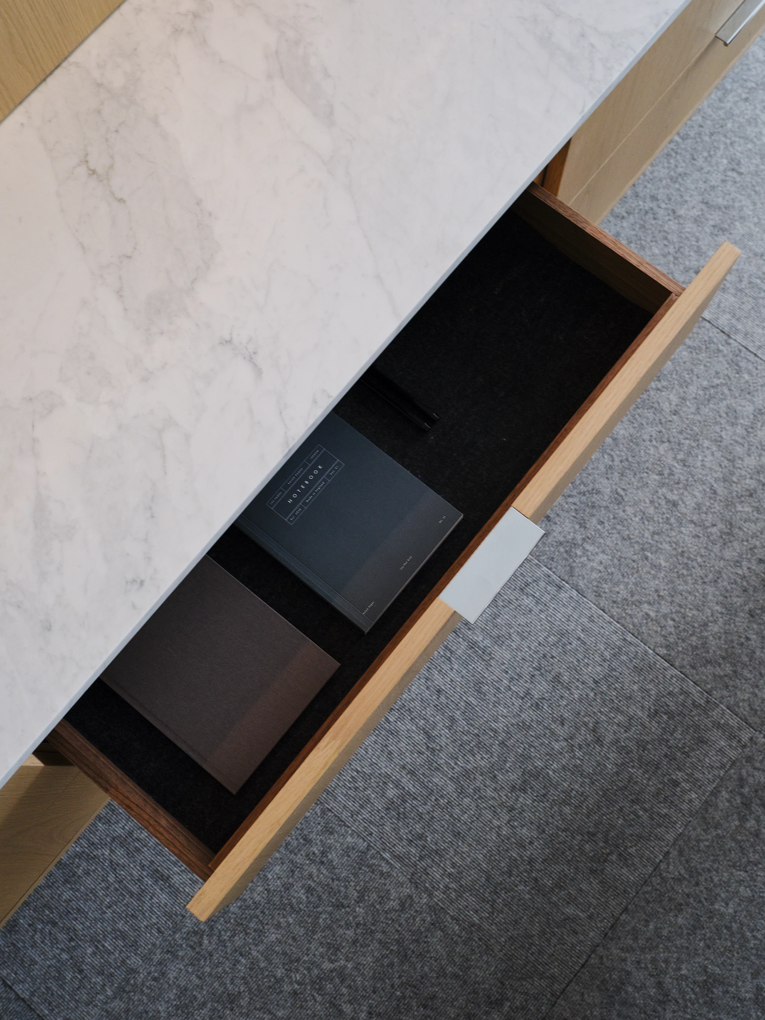 Detail view of a Geiger One Casegoods workwall open storage drawer in Nordic Flat Cut Oak with a White Carrara Marble top, viewed from above in a private office setting.