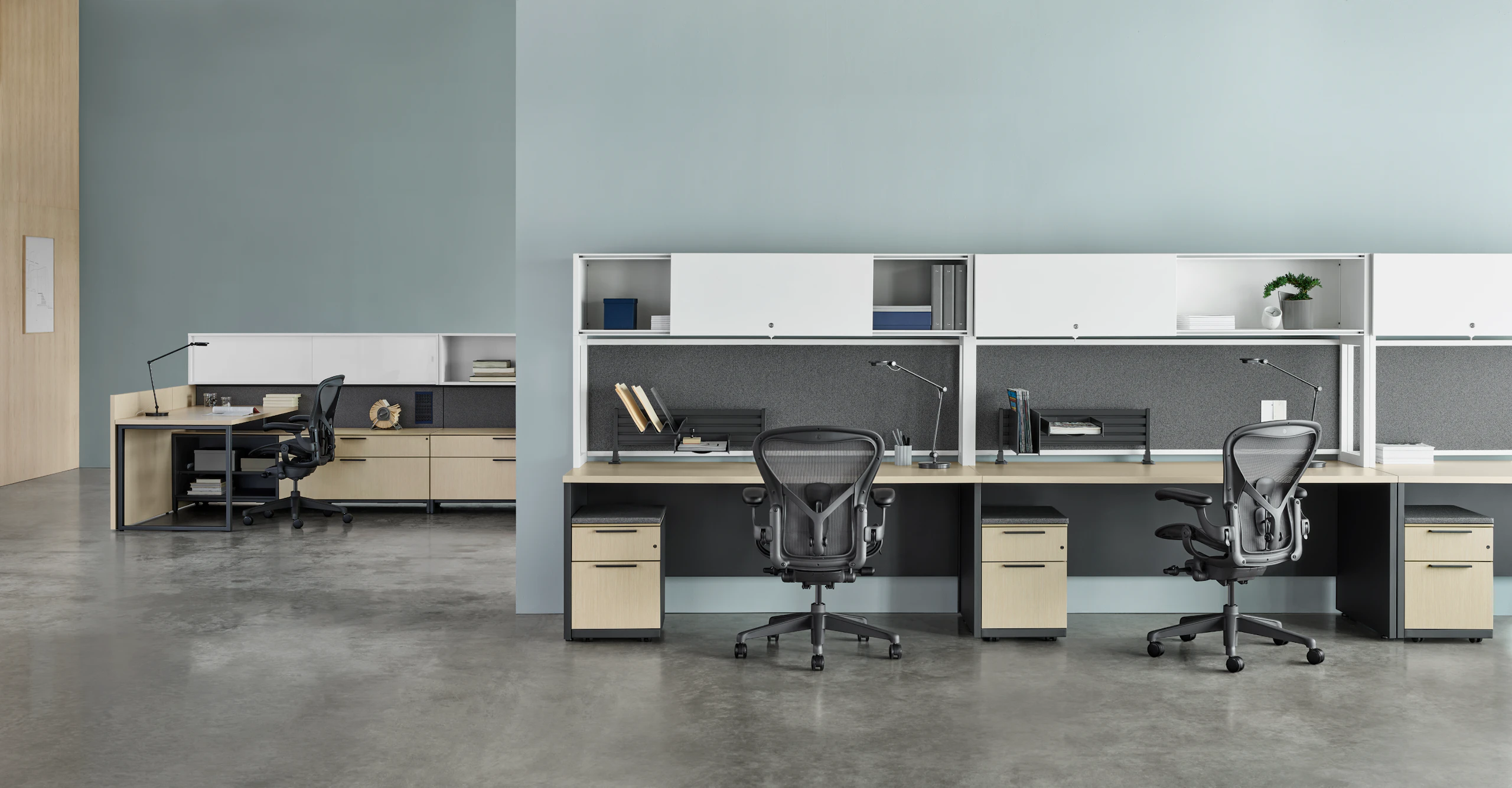 Three Canvas Metal Desks with white upper storage, gray fabric back panels, and light wood surfaces with another workstation in the background.