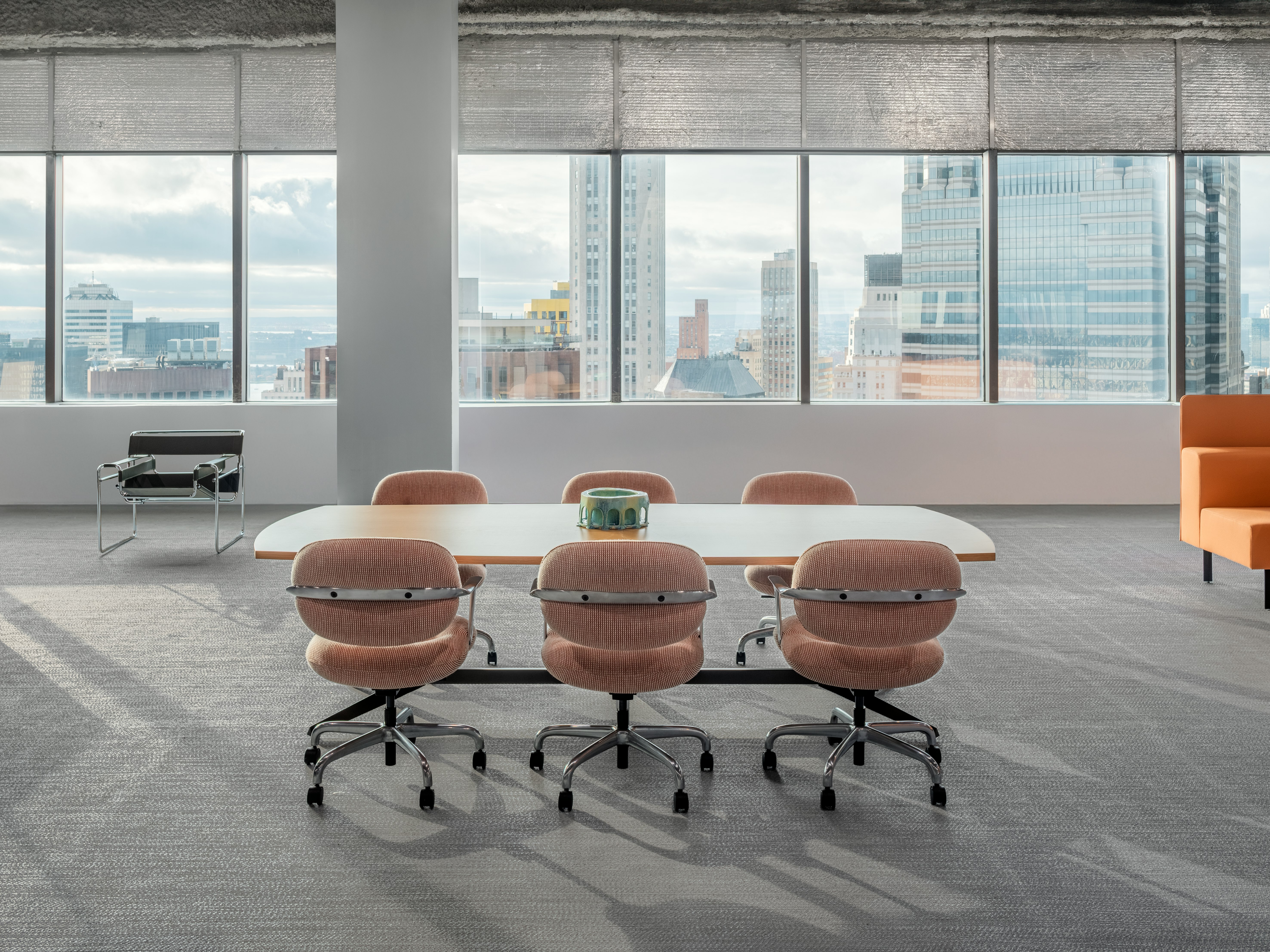 A meeting table and six chairs are in an open office space in front of a large wall of windows. A city skyline is visible through the window.