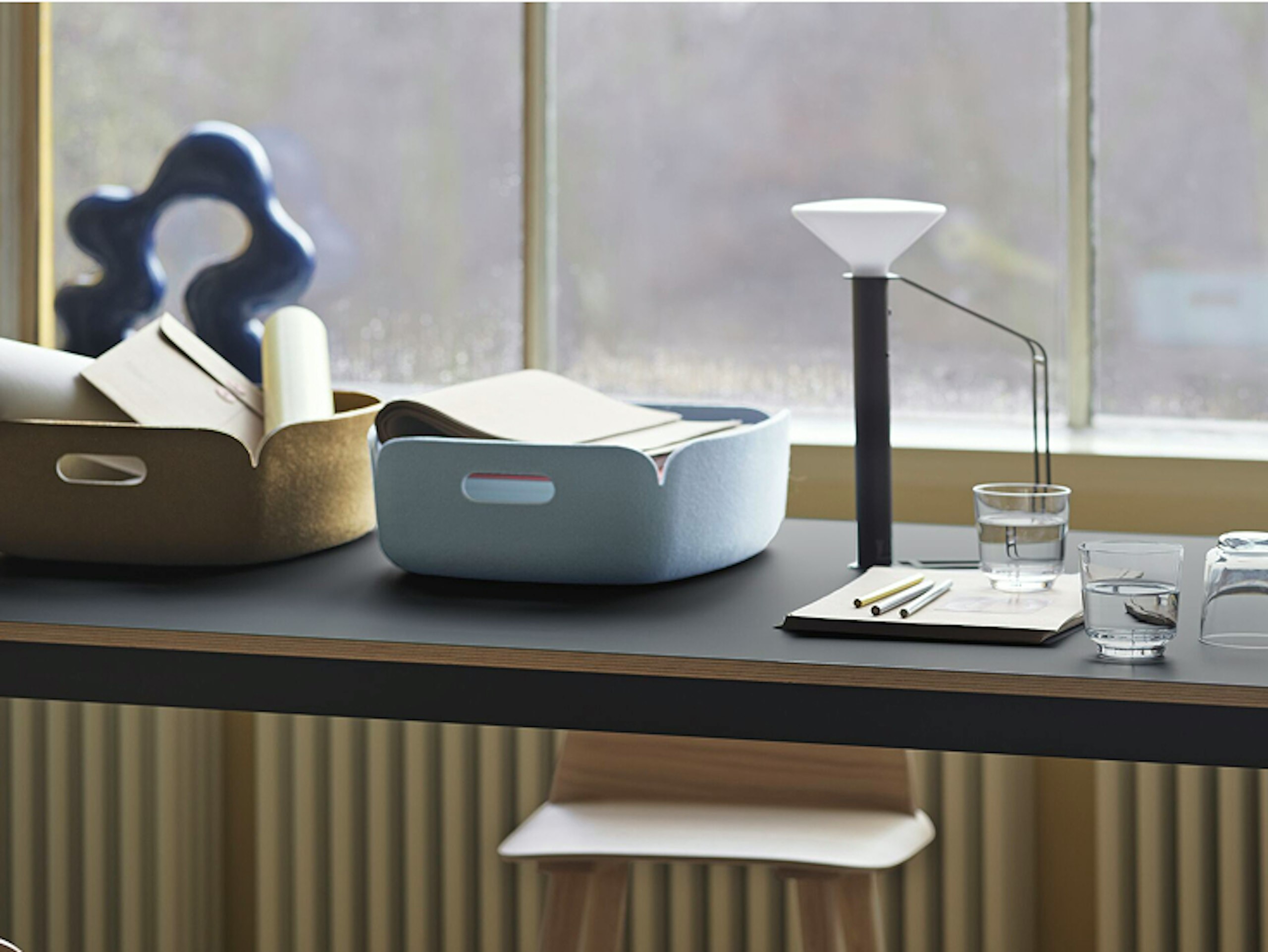 Two Restore Baskets by Muuto, one blue and one yellow, atop a counter height table next to a notepad, lamp, and glasses of water. Sunlight floods through a large window.