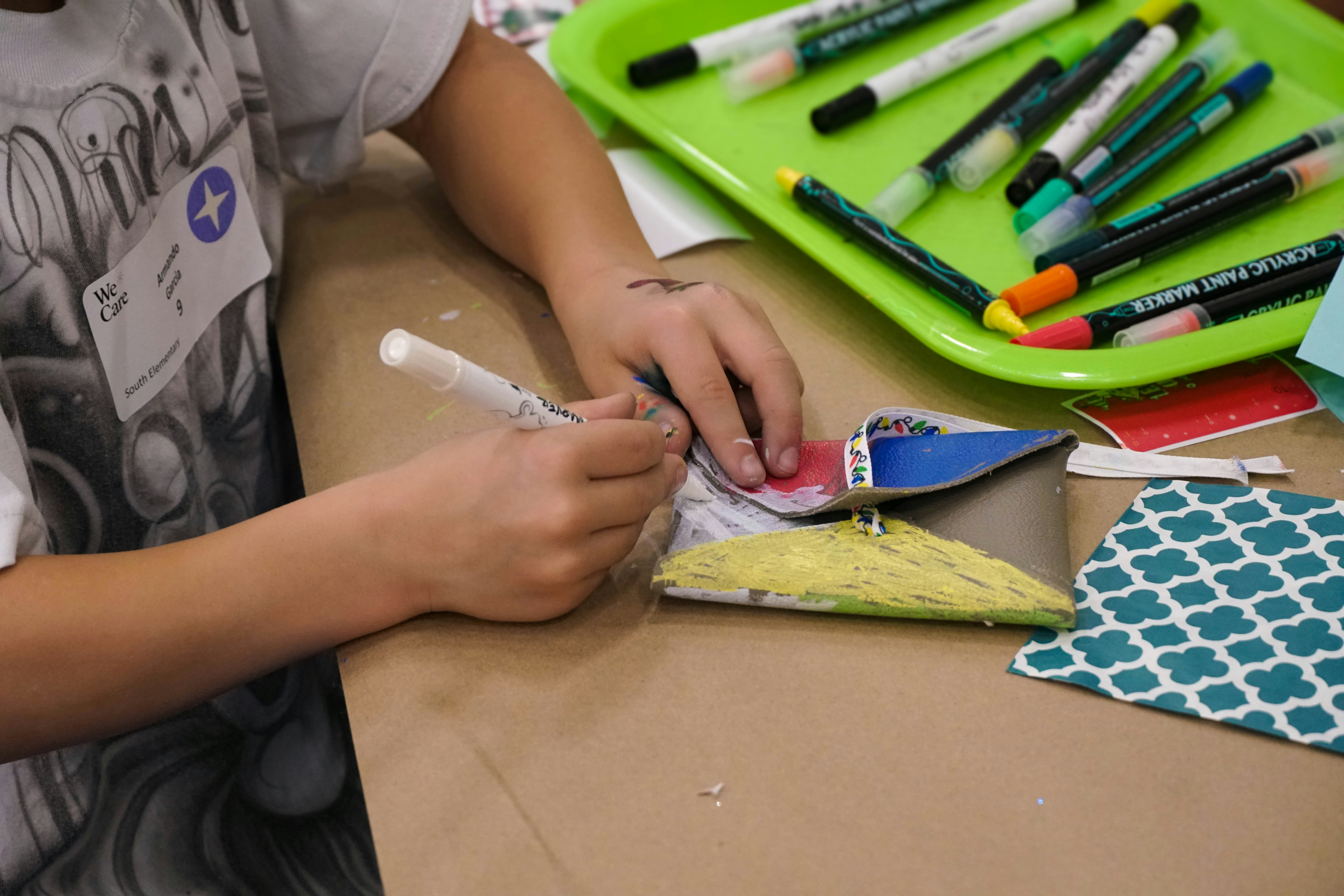 Detail image of a child’s hands drawing on a leather envelope with paint markers at a table with craft materials at a MillerKnoll We Care event, viewed at an angle.