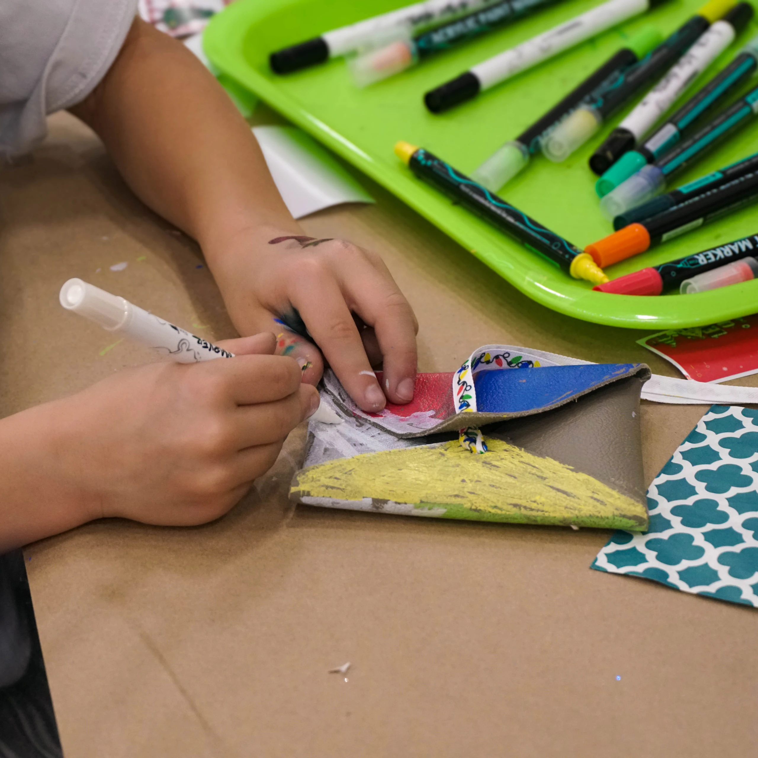 Detail image of a child’s hands drawing on a leather envelope with paint markers at a table with craft materials at a MillerKnoll We Care event, viewed at an angle.