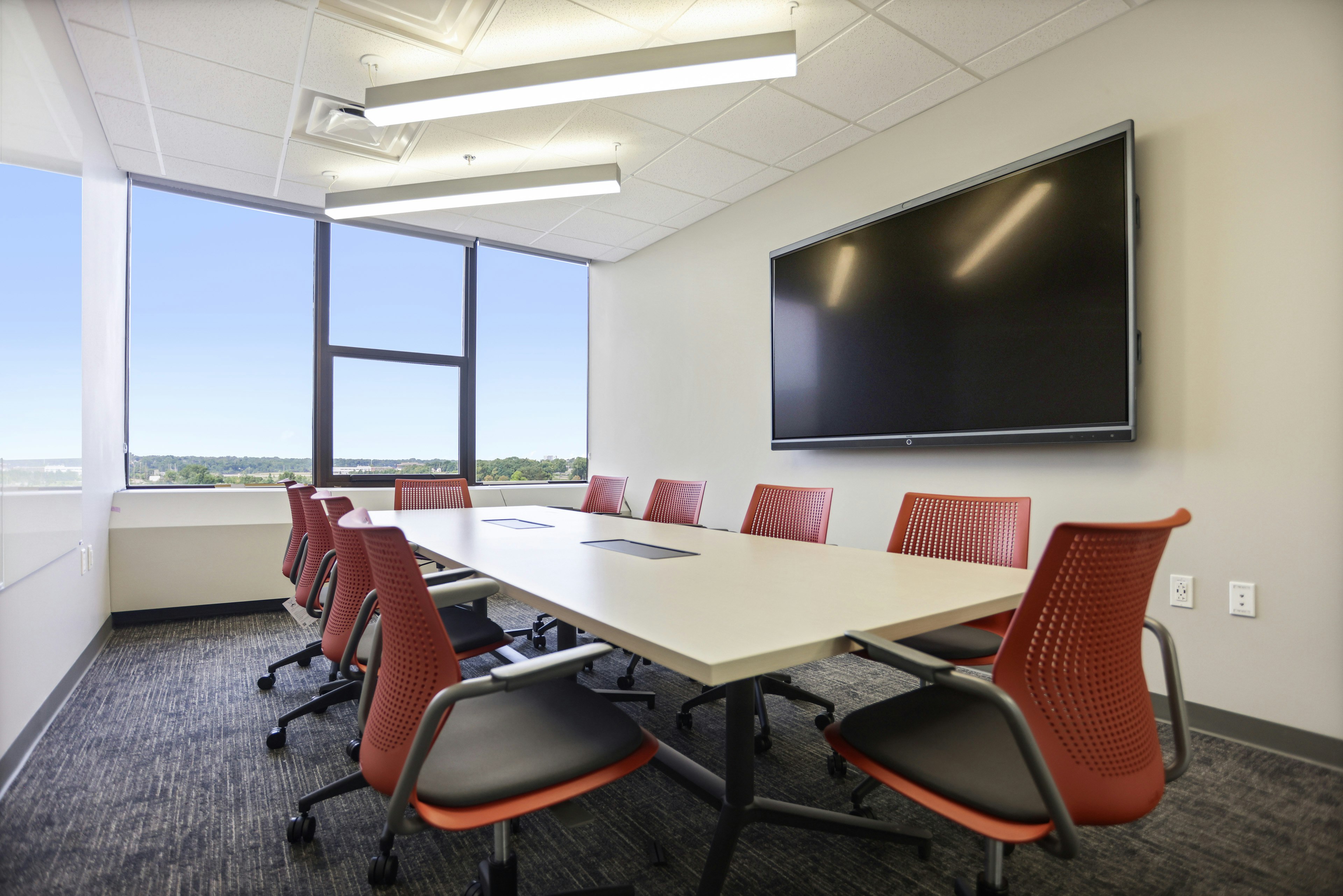 Private conference room furnished with ten red and black Knoll MultiGeneration chairs, a Herman Miller Headway Conference Table, and a large TV mounted on the wall.