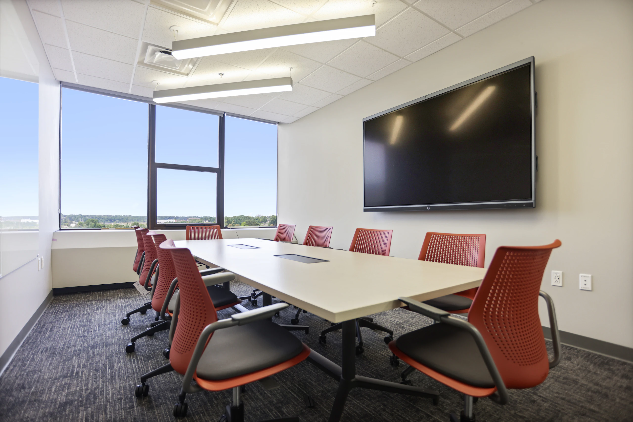 Private conference room furnished with ten red and black Knoll MultiGeneration chairs, a Herman Miller Headway Conference Table, and a large TV mounted on the wall.