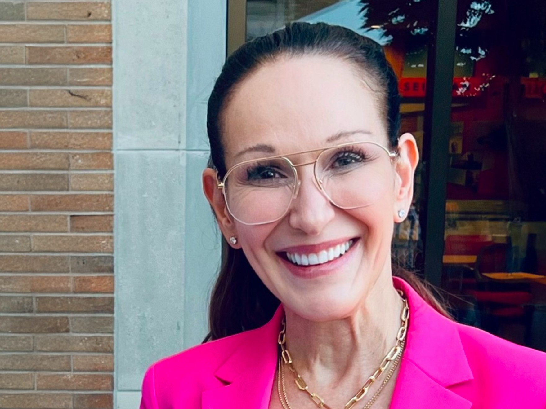 A smiling woman with long brown hair wearing a bright pink blazer, gold necklaces, and glasses.
