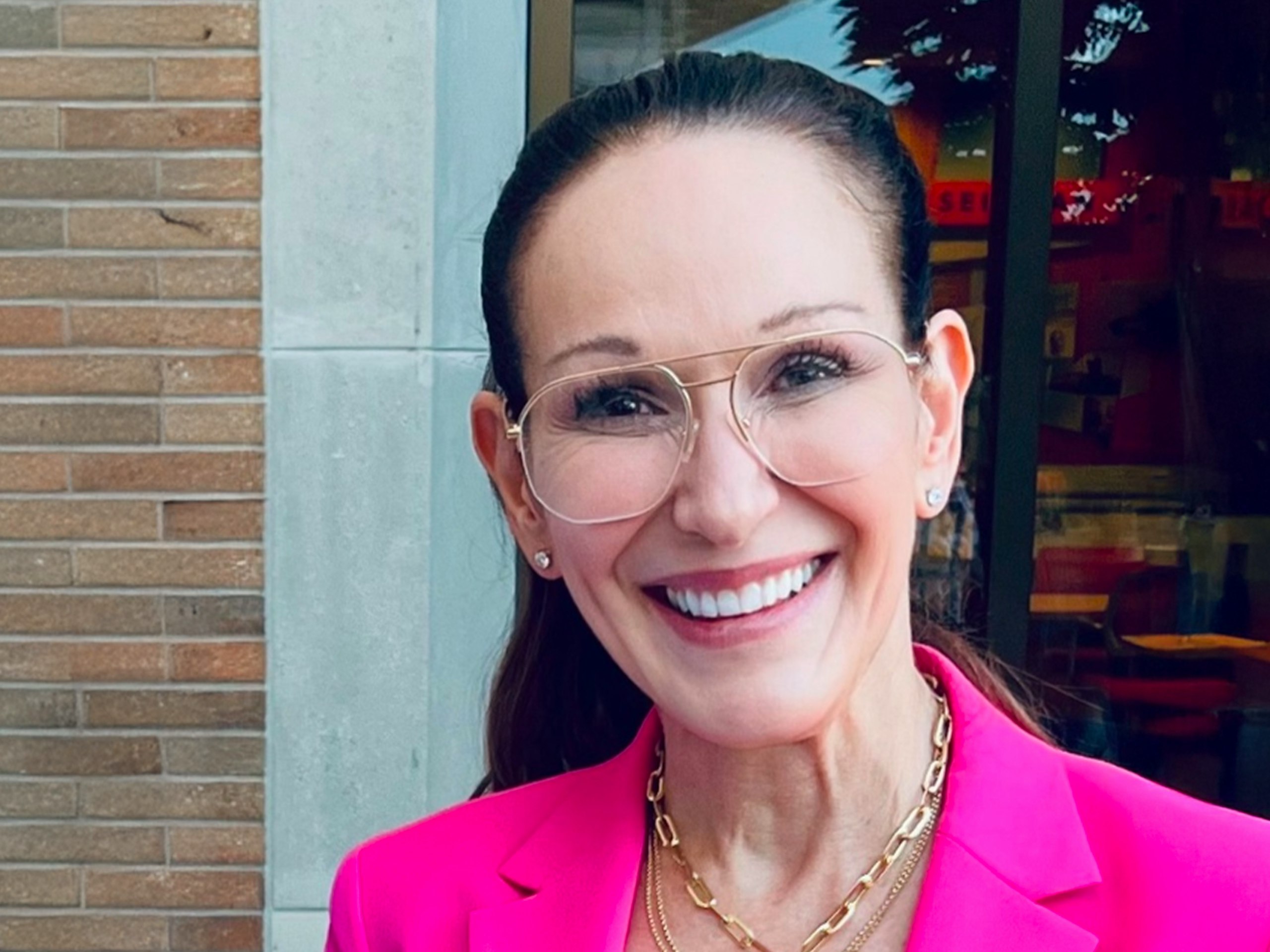 A smiling woman with long brown hair wearing a bright pink blazer, gold necklaces, and glasses.